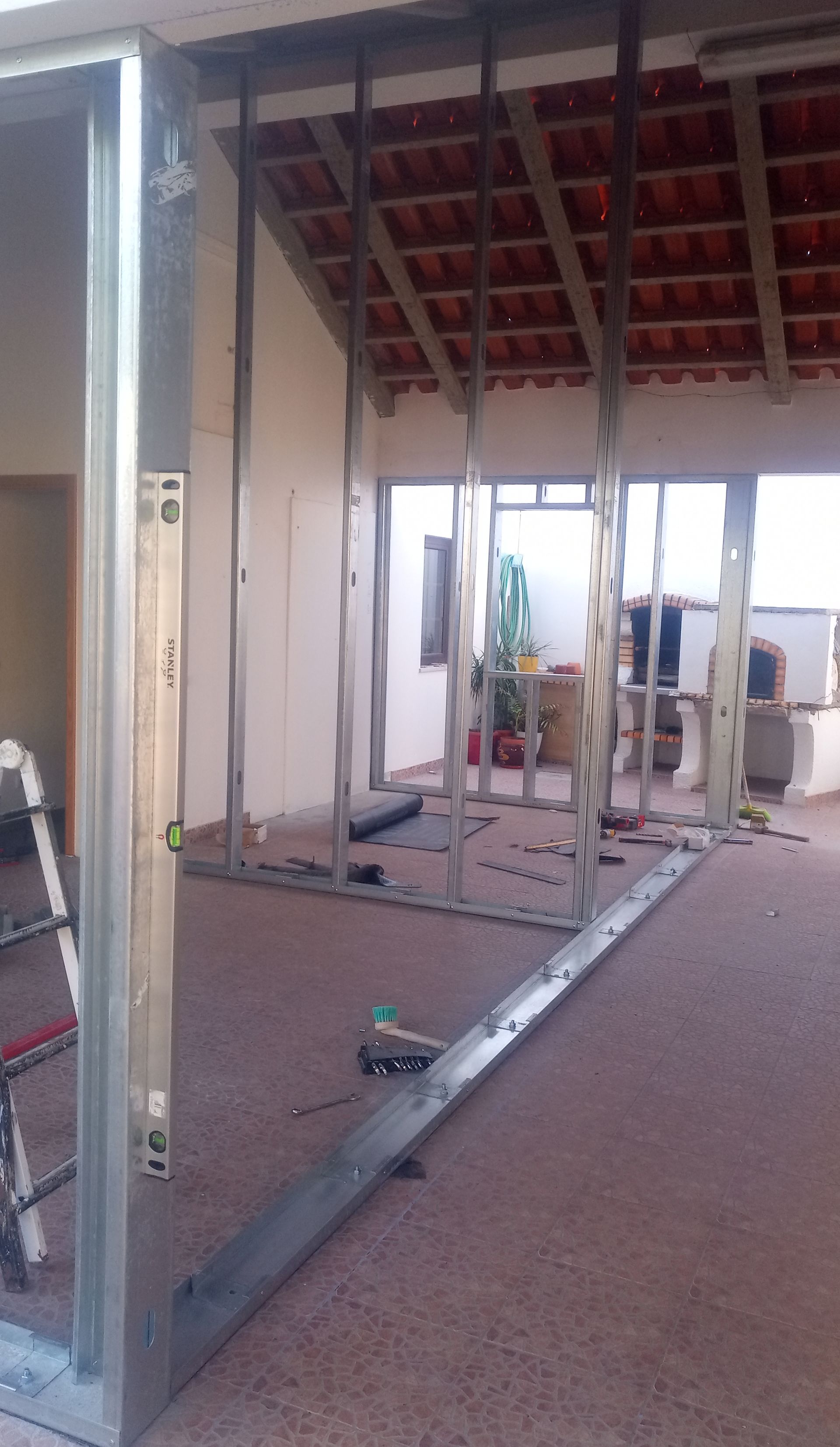 Perfis metálicos delimitam o interior de um cômodo, provavelmente para a construção de uma nova parede. Telhado de telha vermelha visível através da porta.