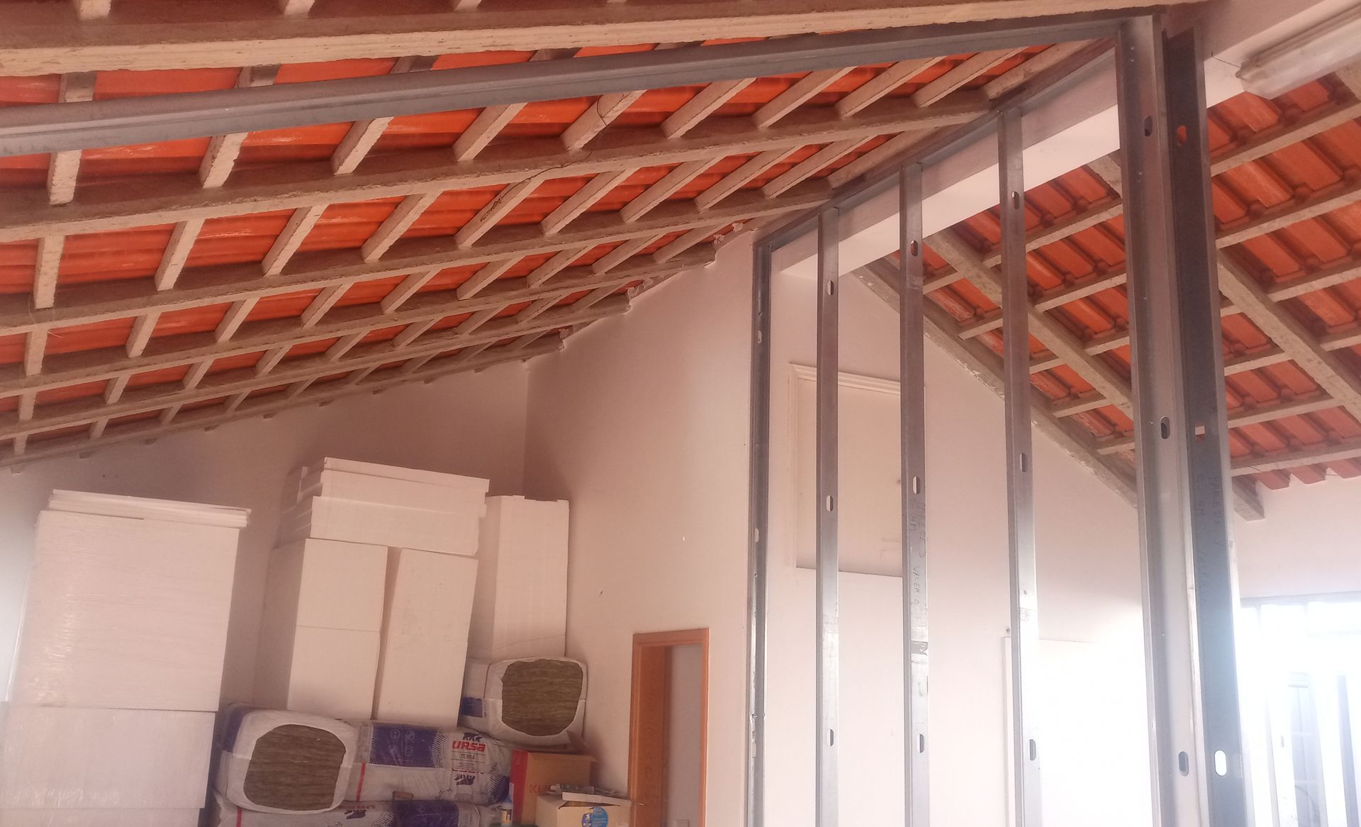 Construção interna mostrando a estrutura metálica da parede, com vigas do telhado visíveis e isolamento empilhado.