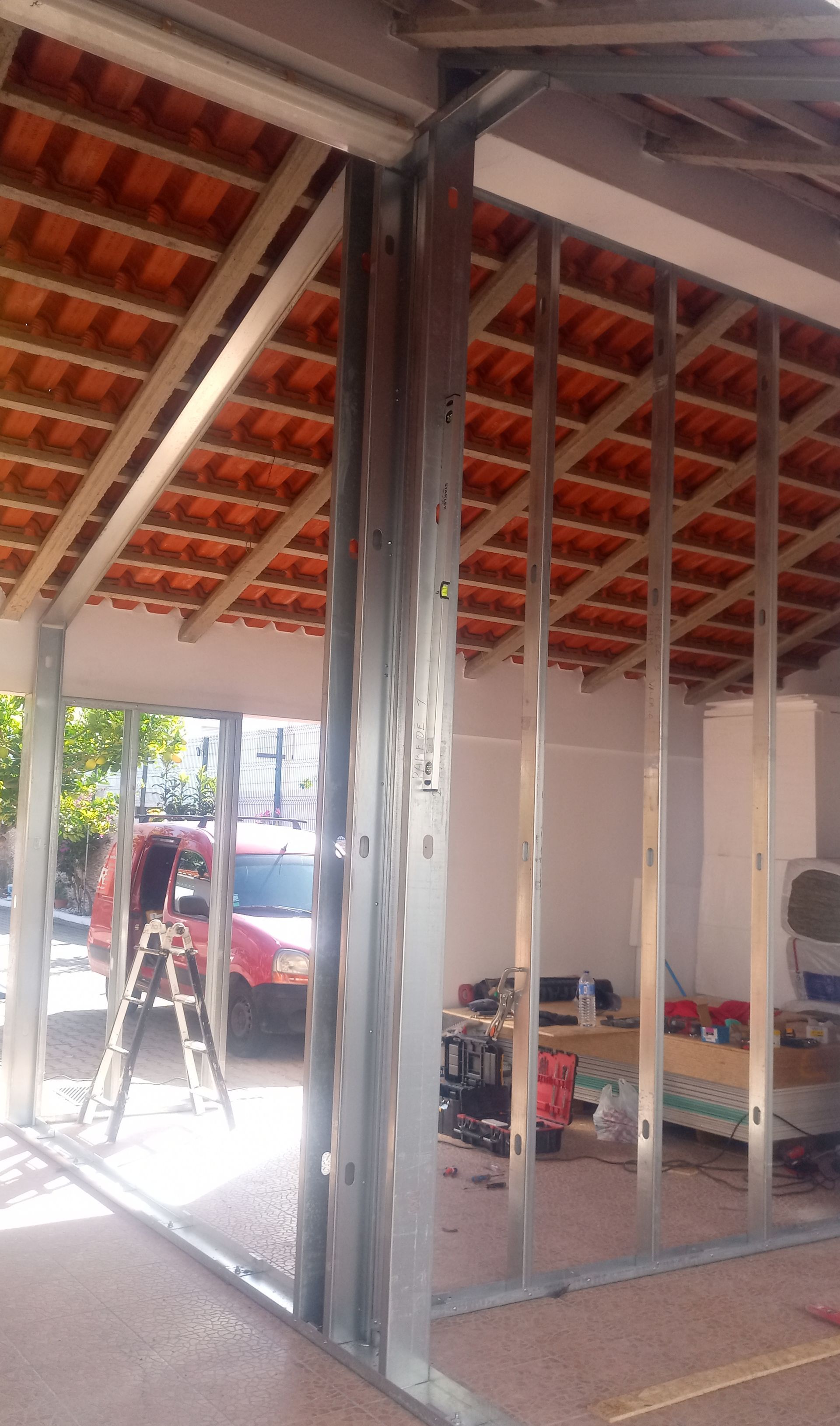 Estrutura metálica para paredes internas sob um telhado de telhas. Um carro vermelho e uma escada são visíveis do lado de fora.