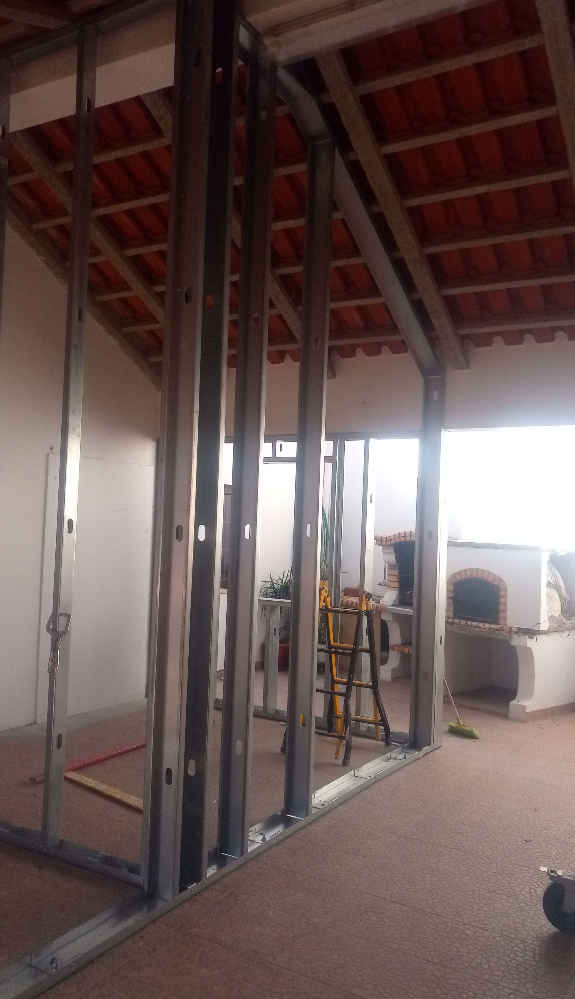 Estrutura de aço para uma parede interna em construção; telhas de terracota visíveis e lareira externa.