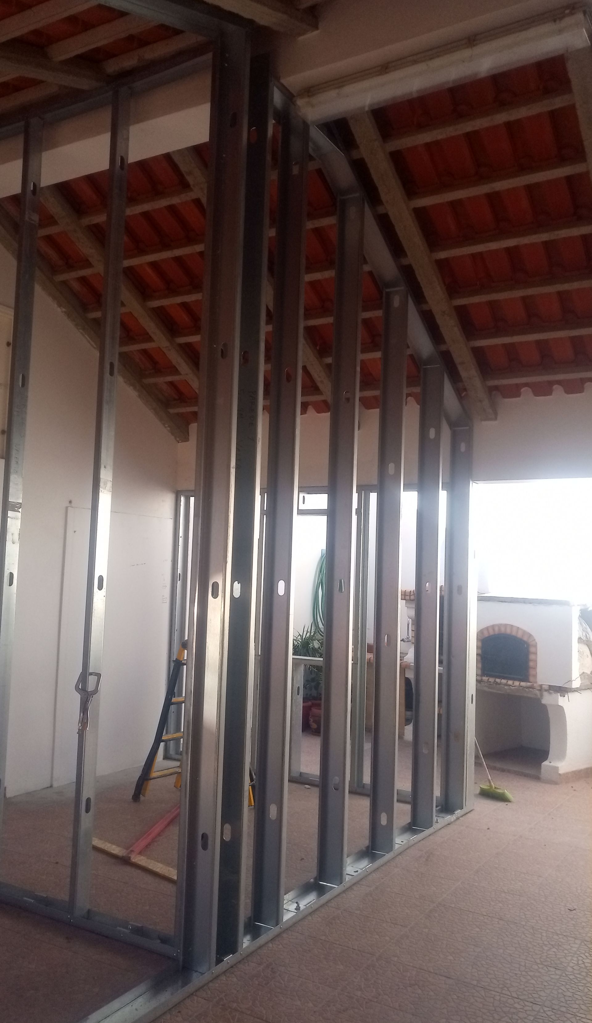 Estrutura metálica formando a armação de uma parede dentro de um edifício em construção, com o telhado de telhas visível.