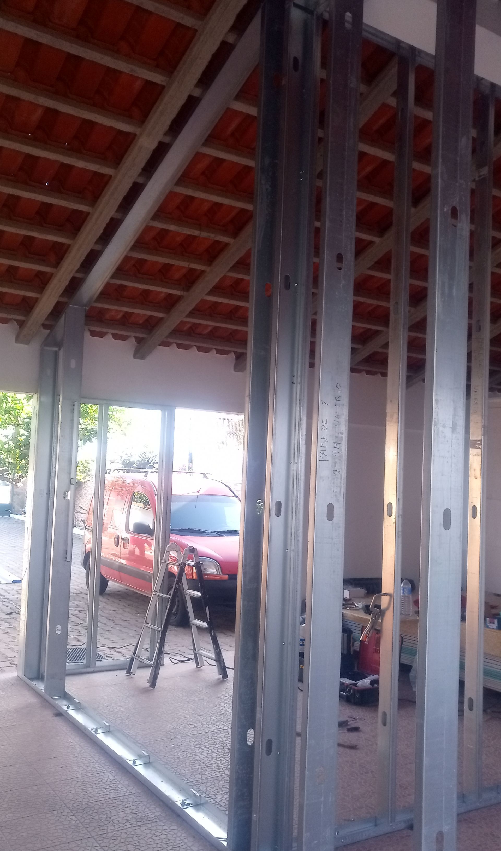 Estrutura metálica de uma parede em construção, revelando o telhado de telha vermelha e a van vermelha.