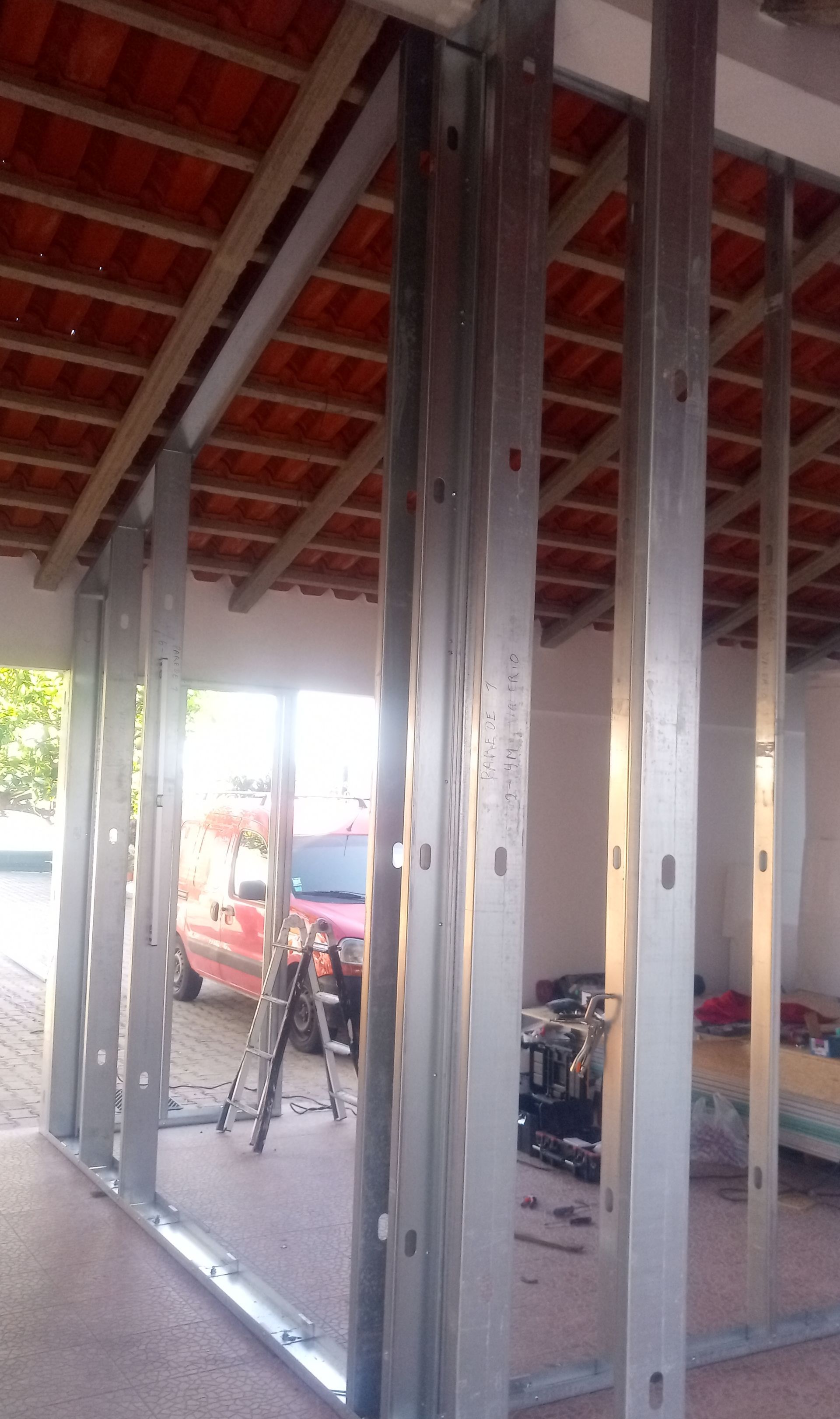 Estrutura metálica para uma parede, no interior de um edifício com telhado de telhas vermelhas e vista para um veículo vermelho.