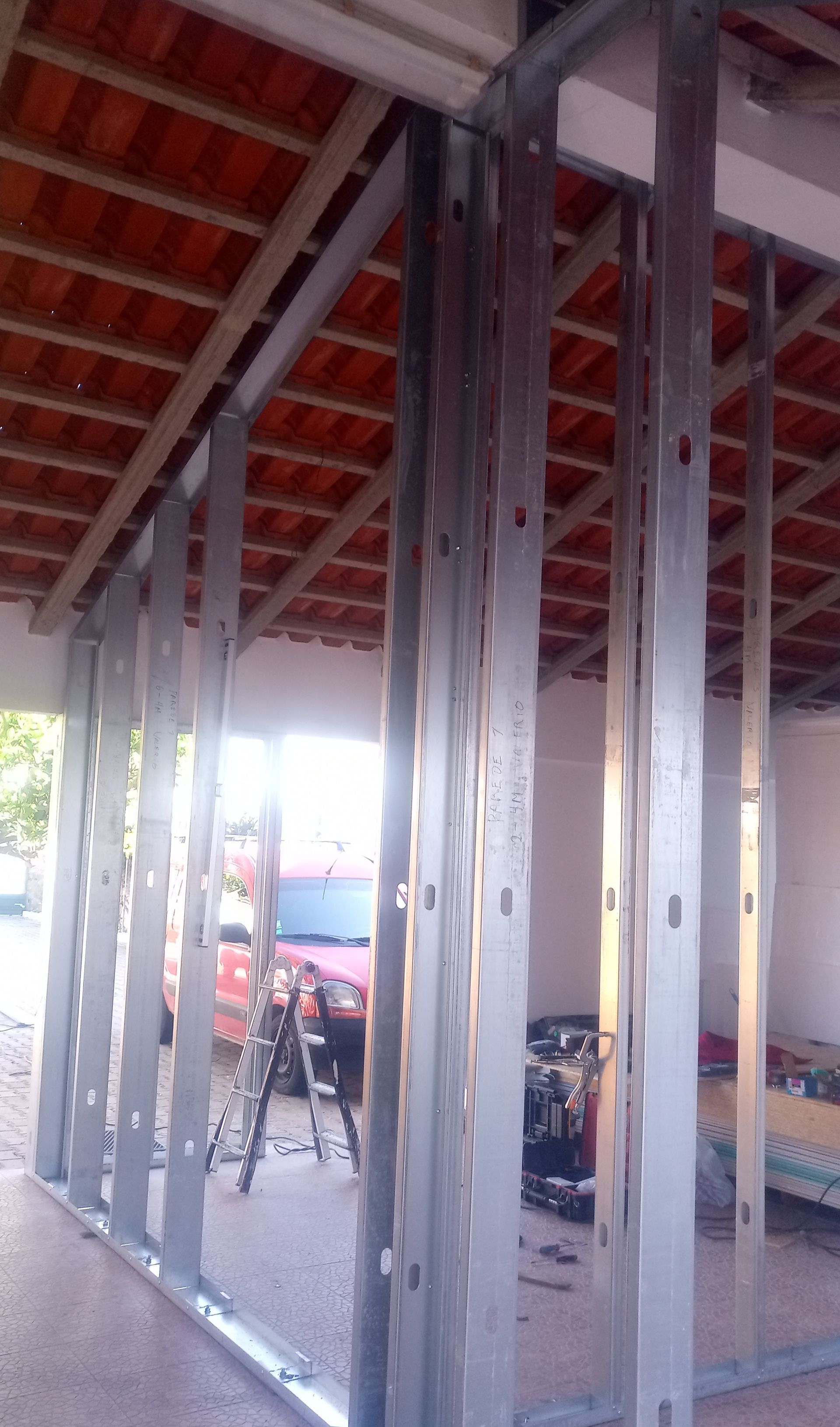 Parede com estrutura metálica em construção, com telhas e ferramentas visíveis ao fundo.