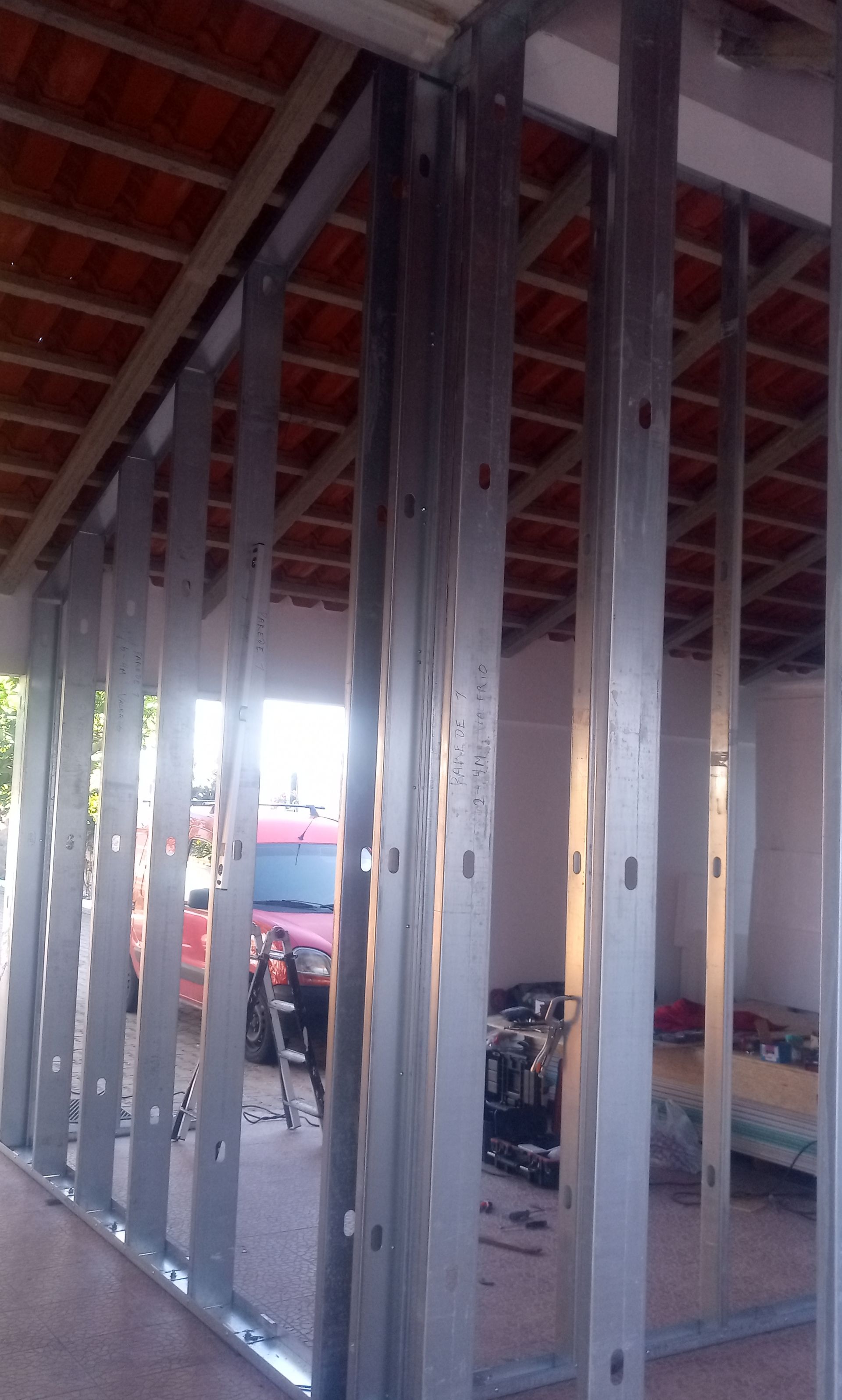 Estrutura metálica de uma parede em construção, com um veículo vermelho visível através da abertura.