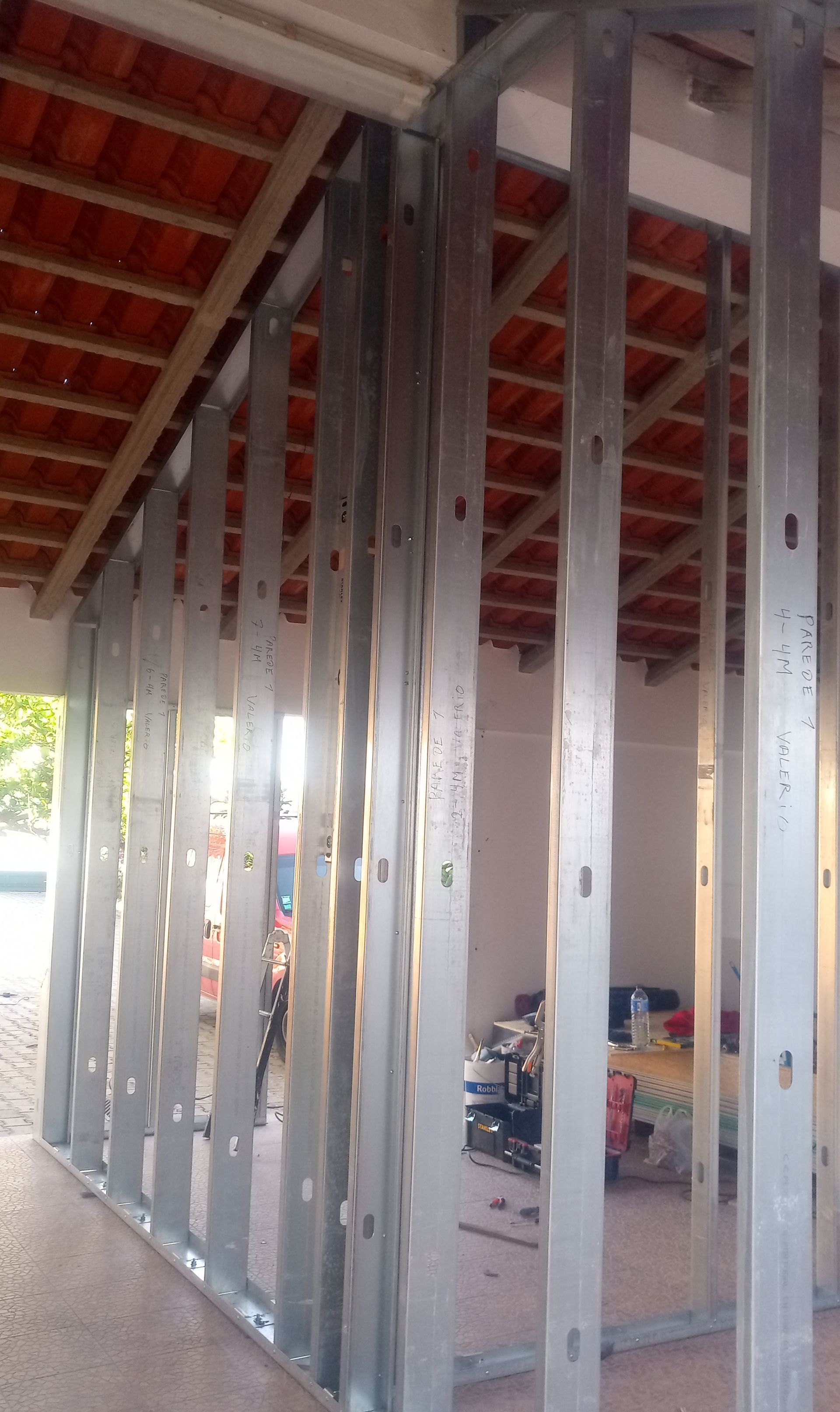 Estrutura metálica para uma parede interna em construção, contra um telhado de telha vermelha e paredes inacabadas.