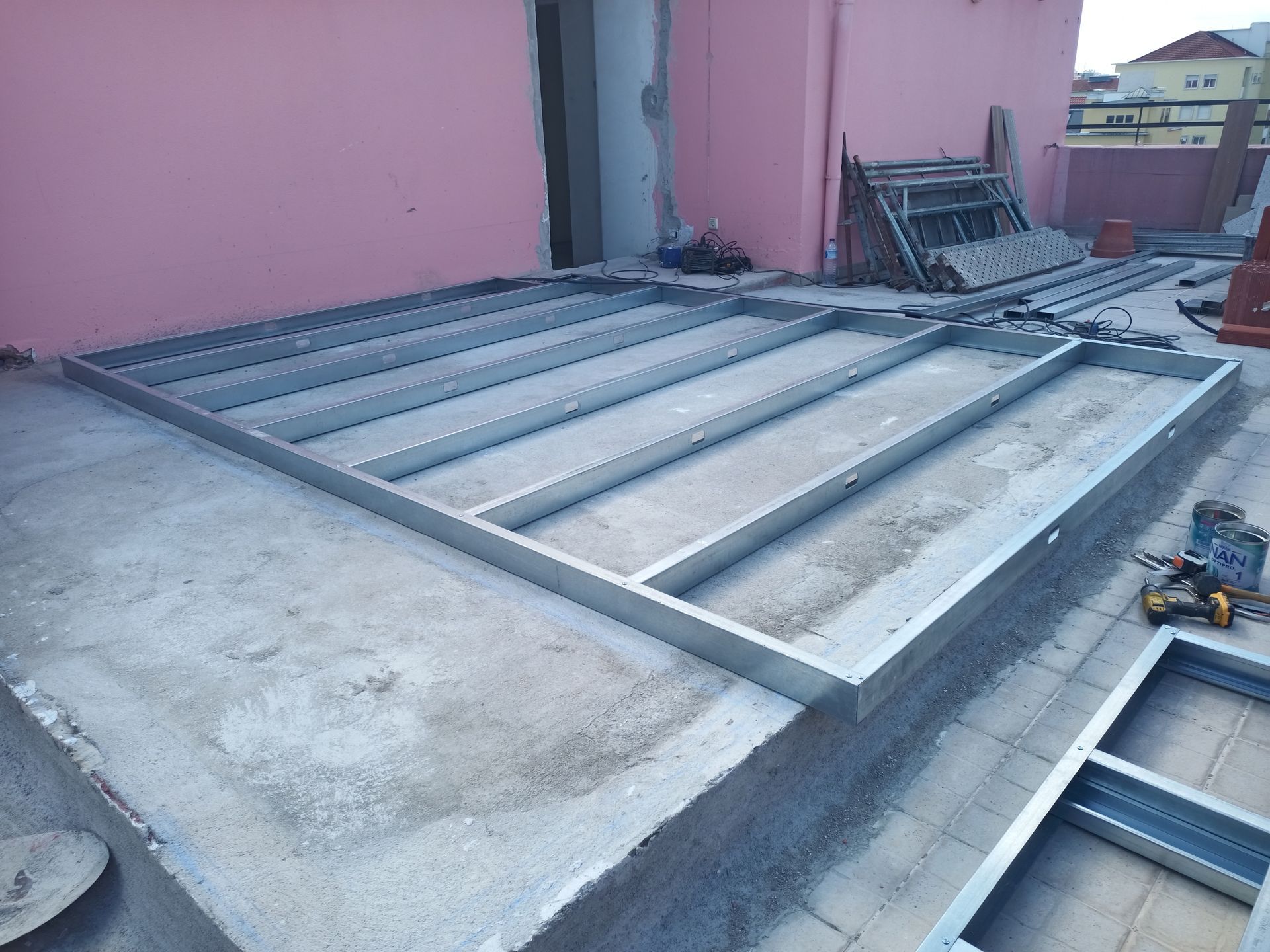 Estrutura metálica assentada sobre uma superfície de concreto, provavelmente para construção em um telhado. Parede rosa ao fundo.