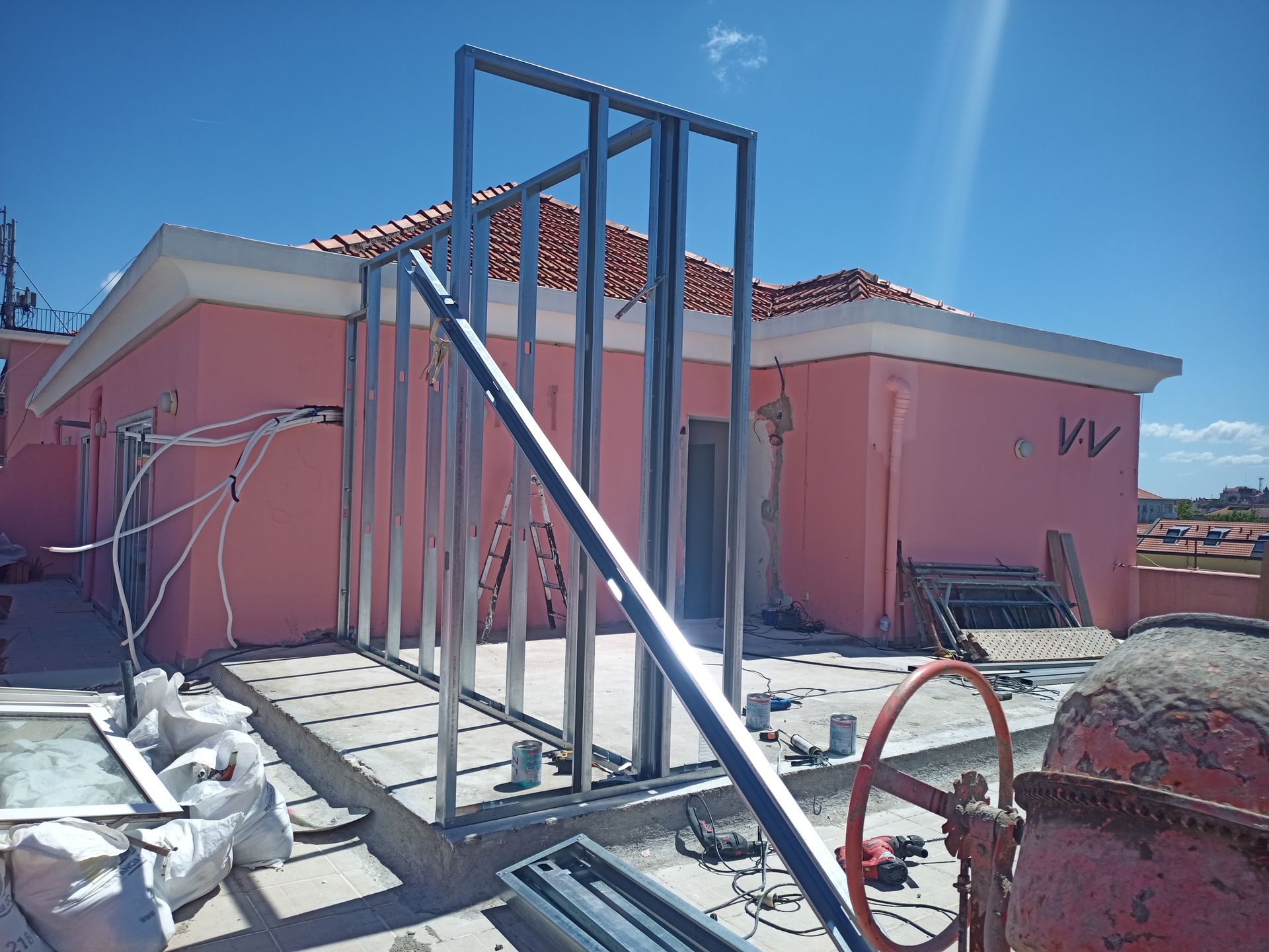 Canteiro de obras com estrutura metálica encostada em um prédio rosa. Céu azul, piso de cimento, equipamentos visíveis.