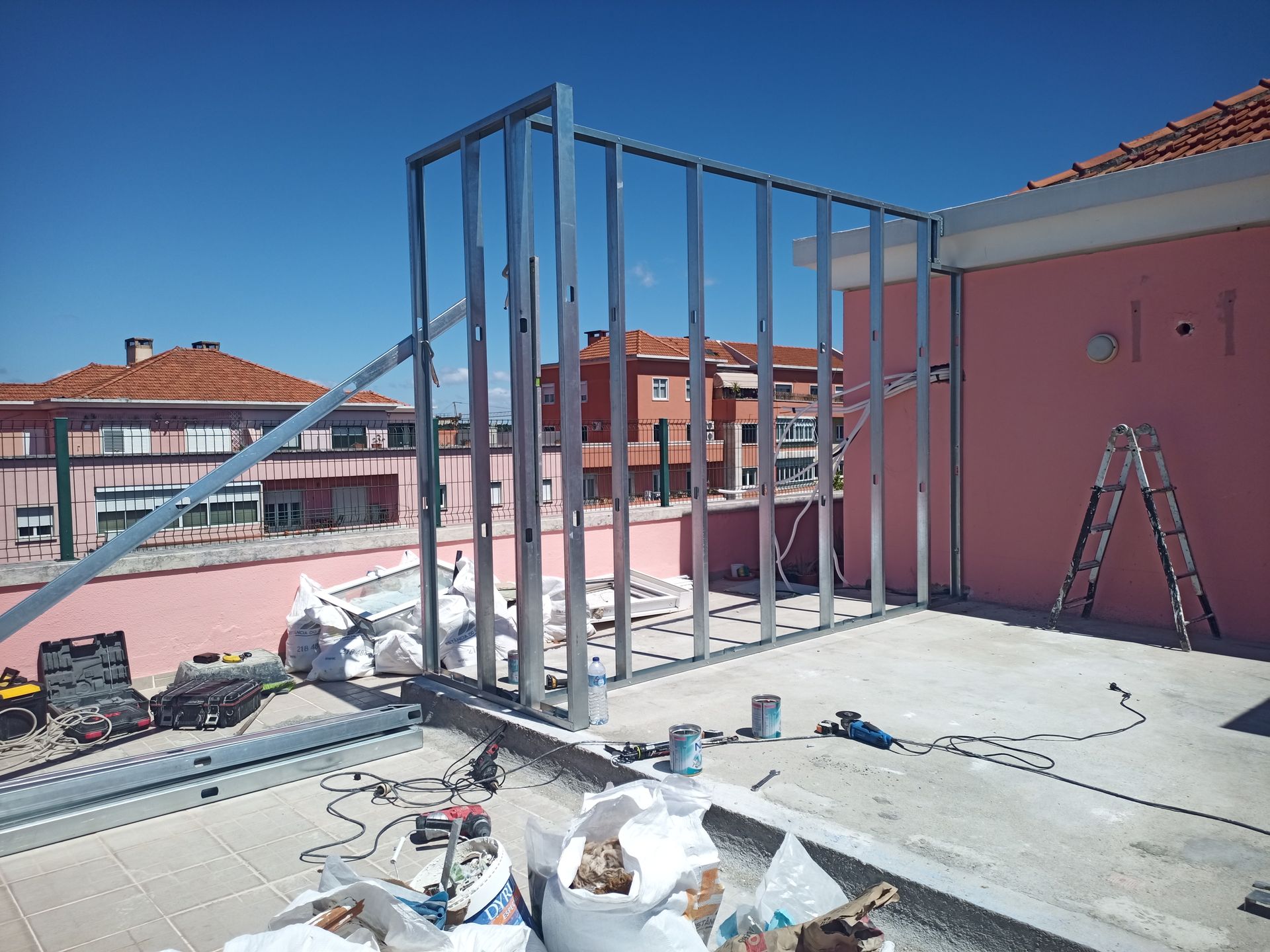 Estrutura metálica para uma parede em construção em um telhado, com um prédio rosa e céu limpo visíveis.