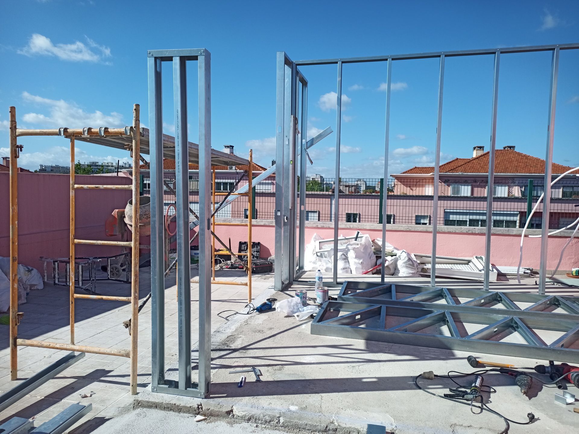 Canteiro de obras com estrutura de aço. Andaimes e materiais em um telhado com prédios ao fundo.