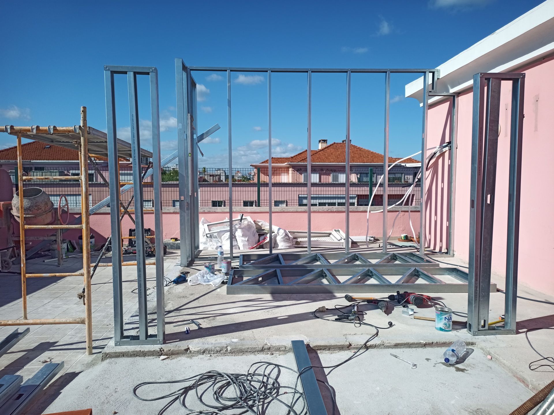 Estrutura de aço que sustenta uma parede sobre uma superfície de concreto. Prédios e céu ao fundo.