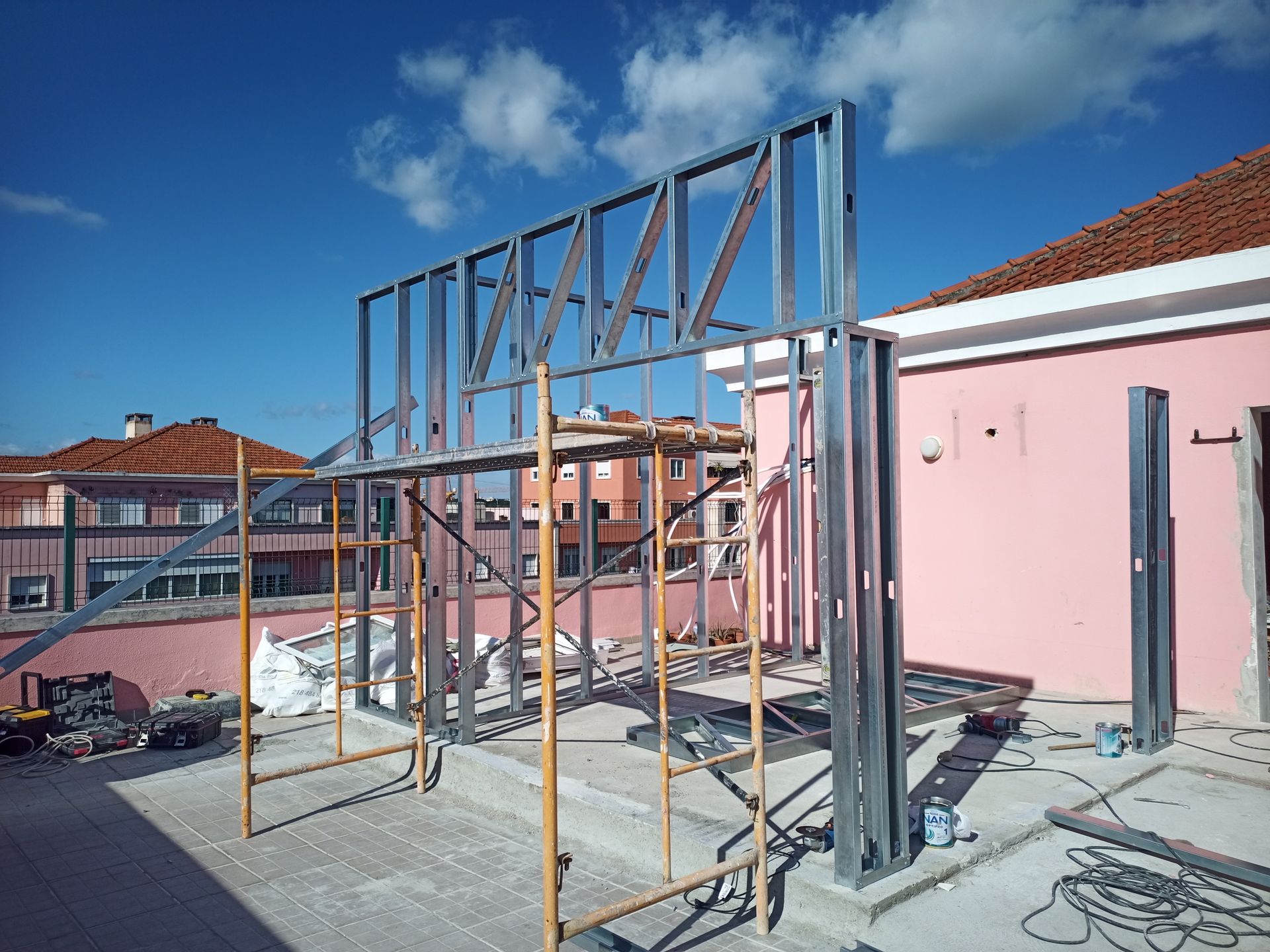 Estrutura metálica em construção sobre um telhado, com andaimes, tendo como pano de fundo um prédio rosa e um céu azul.