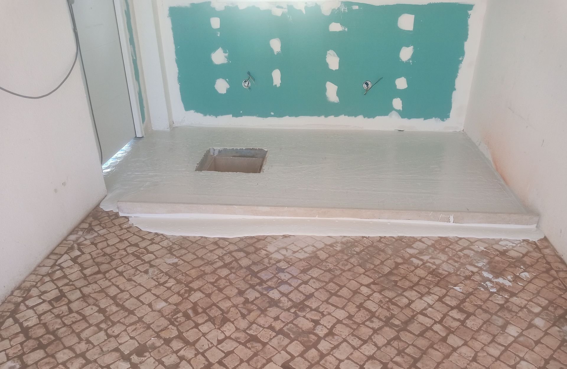 Banheiro em construção, mostrando uma área de chuveiro com plataforma elevada, paredes inacabadas e piso de mosaico.