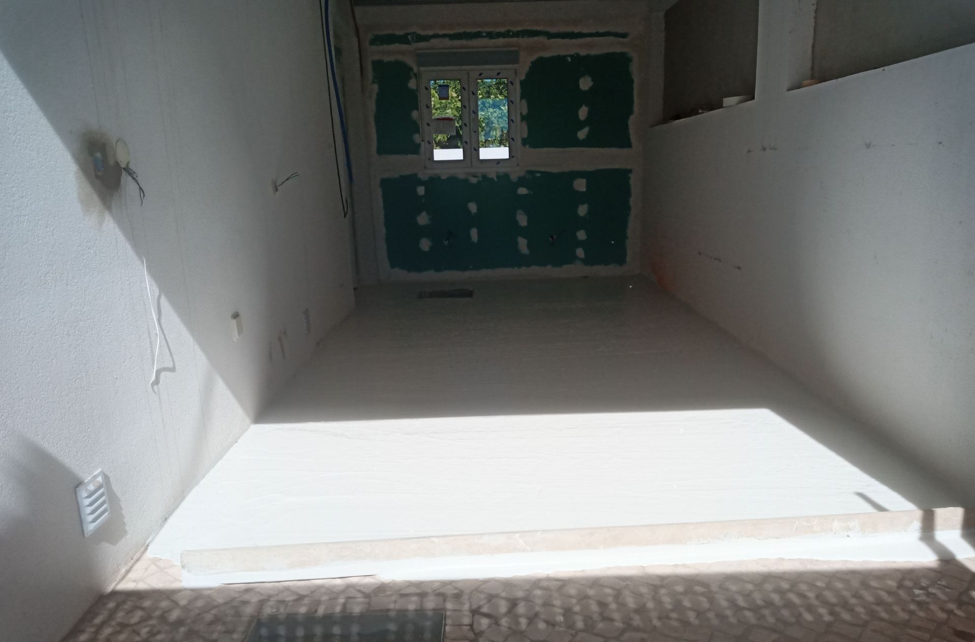 Vista interior de um cômodo em construção com paredes brancas, uma janela e estrutura aparente.