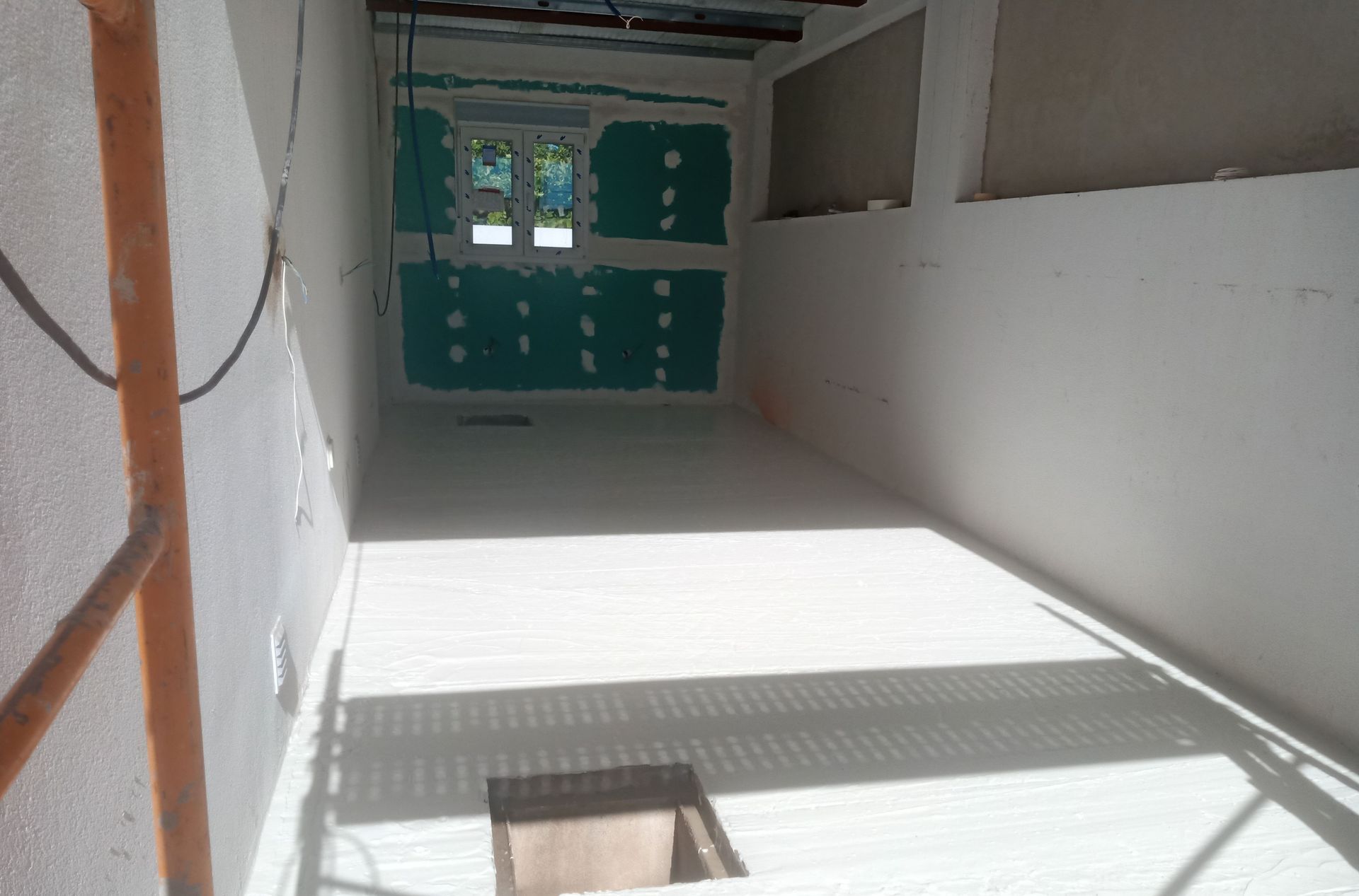 Interior de uma sala em construção, com paredes e piso brancos. Andaimes à esquerda.