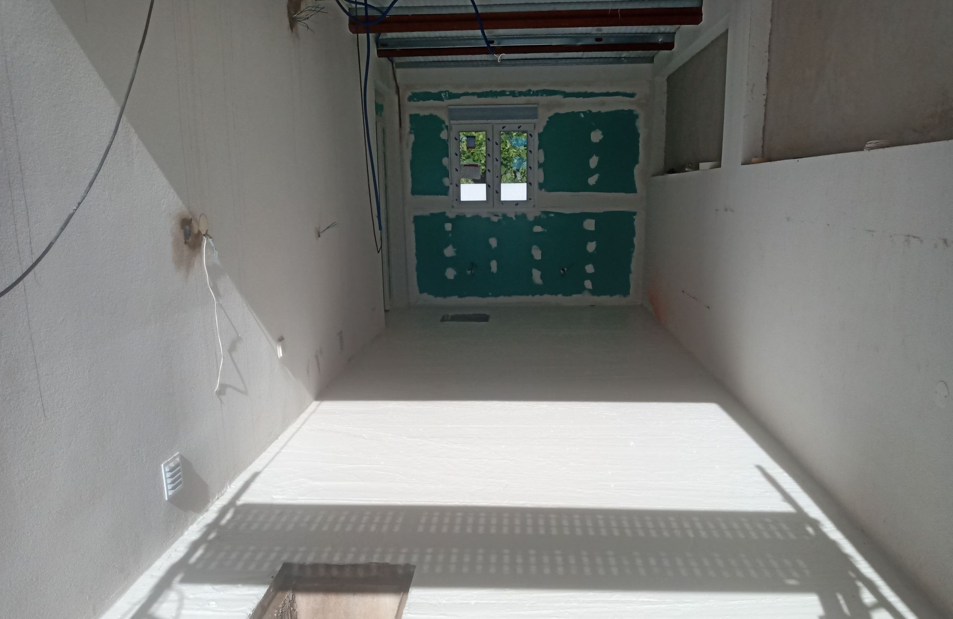 Quarto interior em construção, com drywall verde visível ao redor de uma janela e paredes brancas.