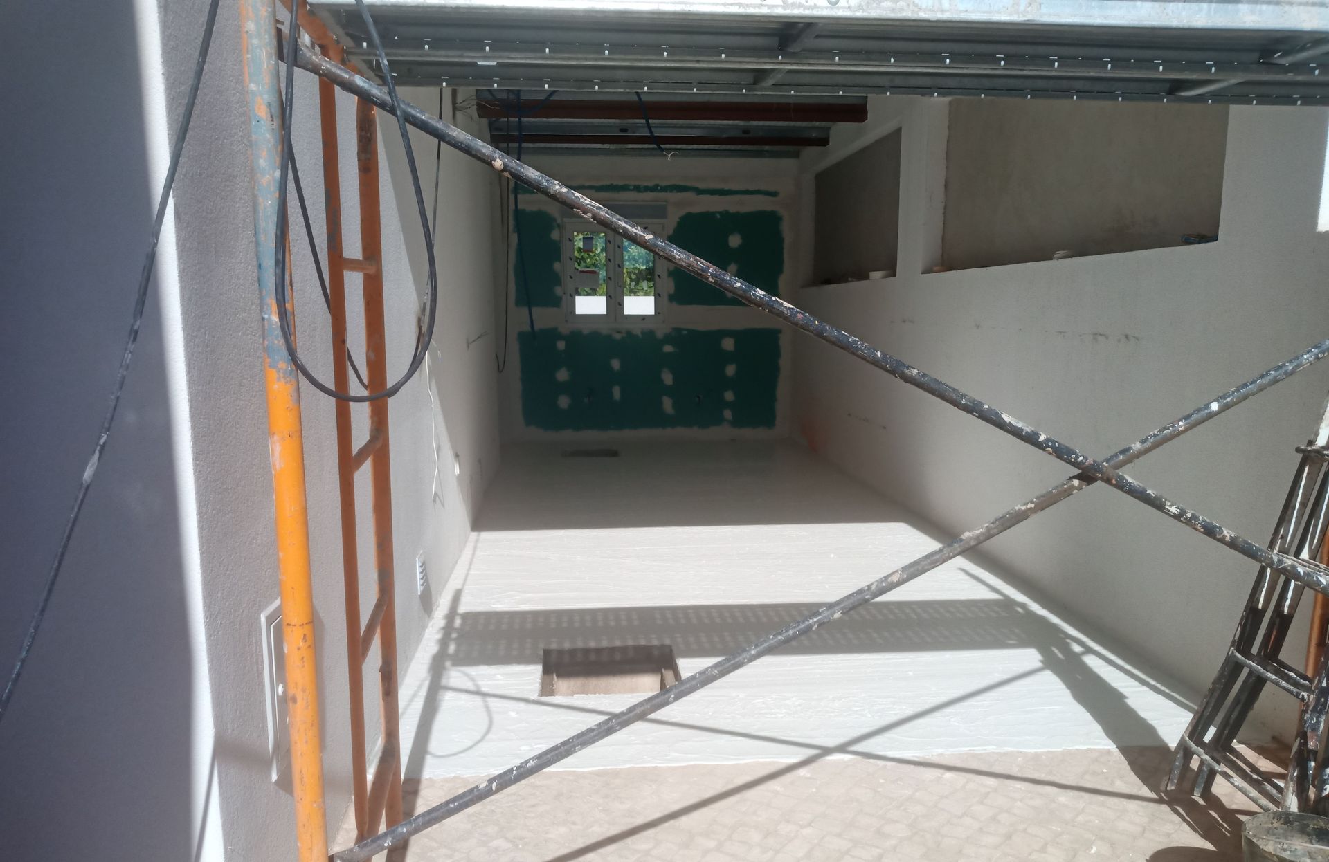 Construção de um pequeno cômodo. Andaimes e vigas metálicas laranja são visíveis. Paredes e piso brancos.
