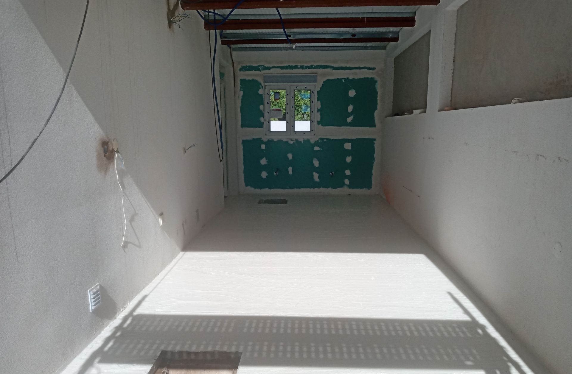 Interior de um cômodo em construção, com paredes brancas, uma janela e vigas expostas no teto.