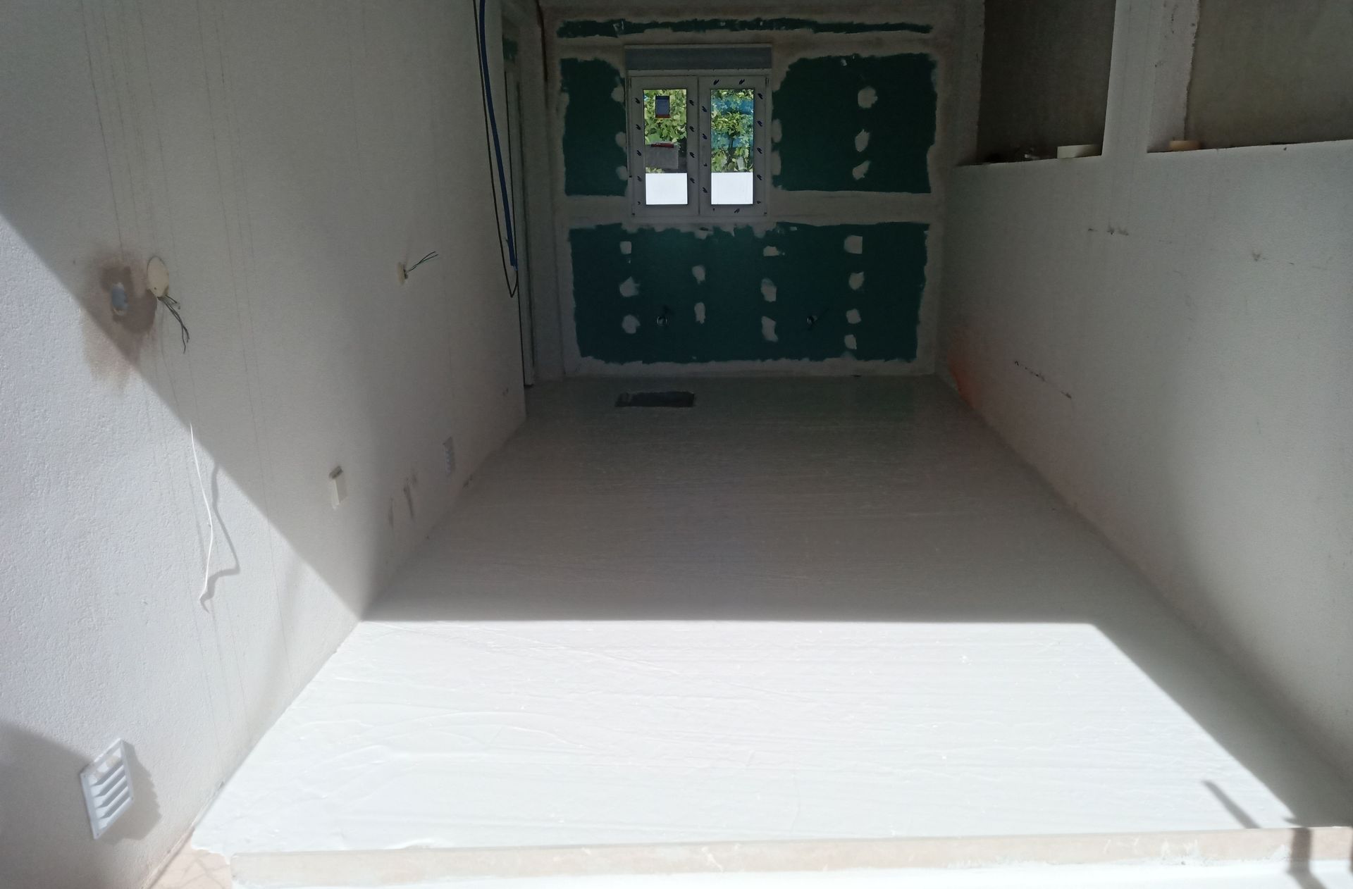 Interior de um cômodo em construção, com paredes brancas e uma janela ao fundo; o drywall ao redor da janela é visível.