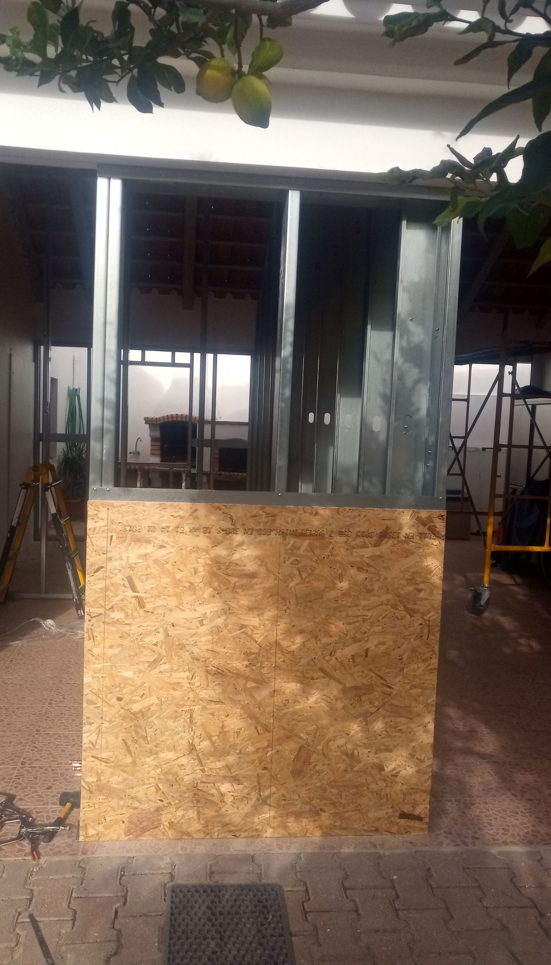 Canteiro de obras: painel de madeira em frente a uma estrutura metálica, ambiente externo com andaimes e árvore.