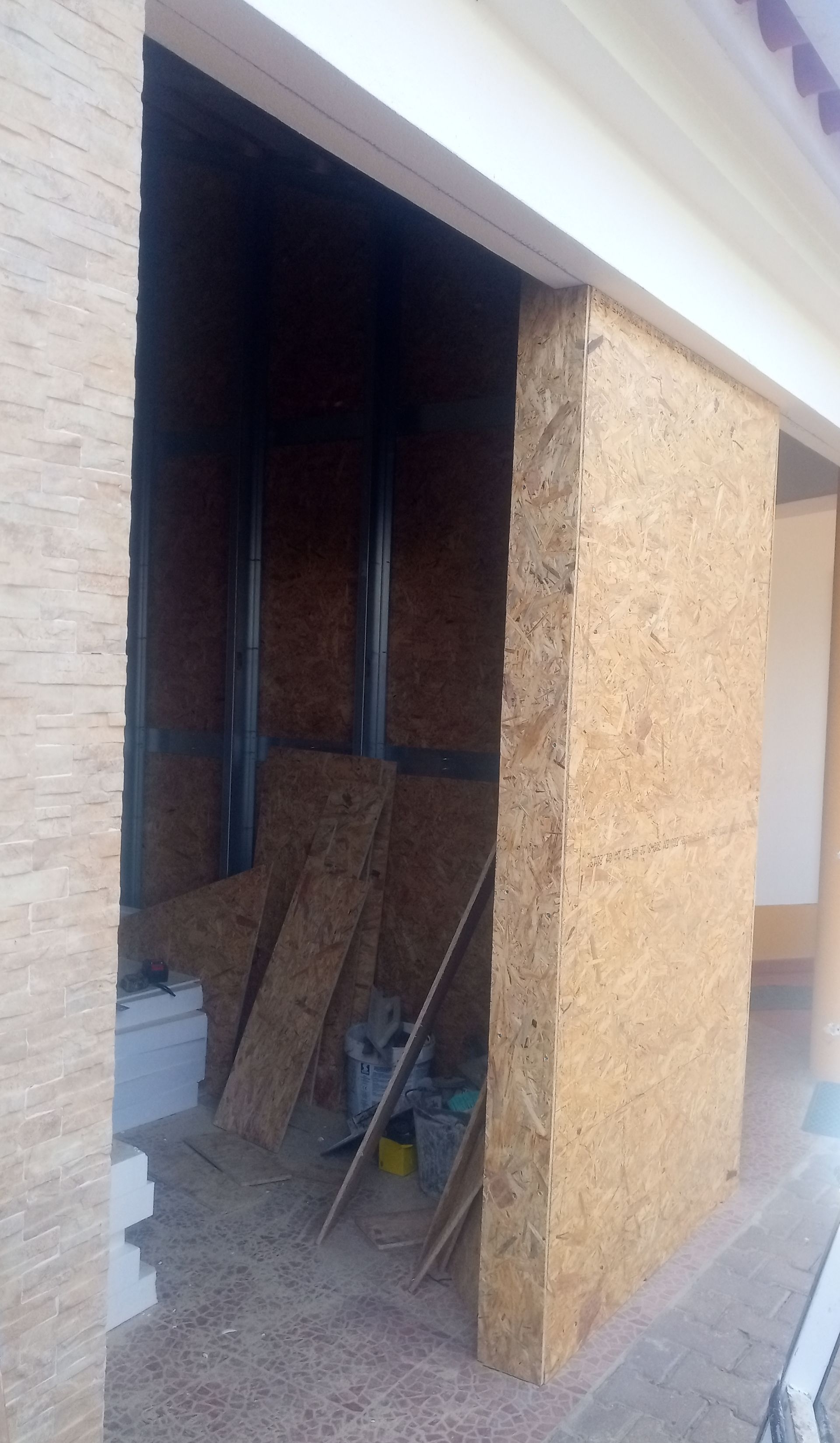 Canteiro de obras externo: revestimento de placas OSB e porta parcialmente emoldurada. Tons de marrom, bege e branco.