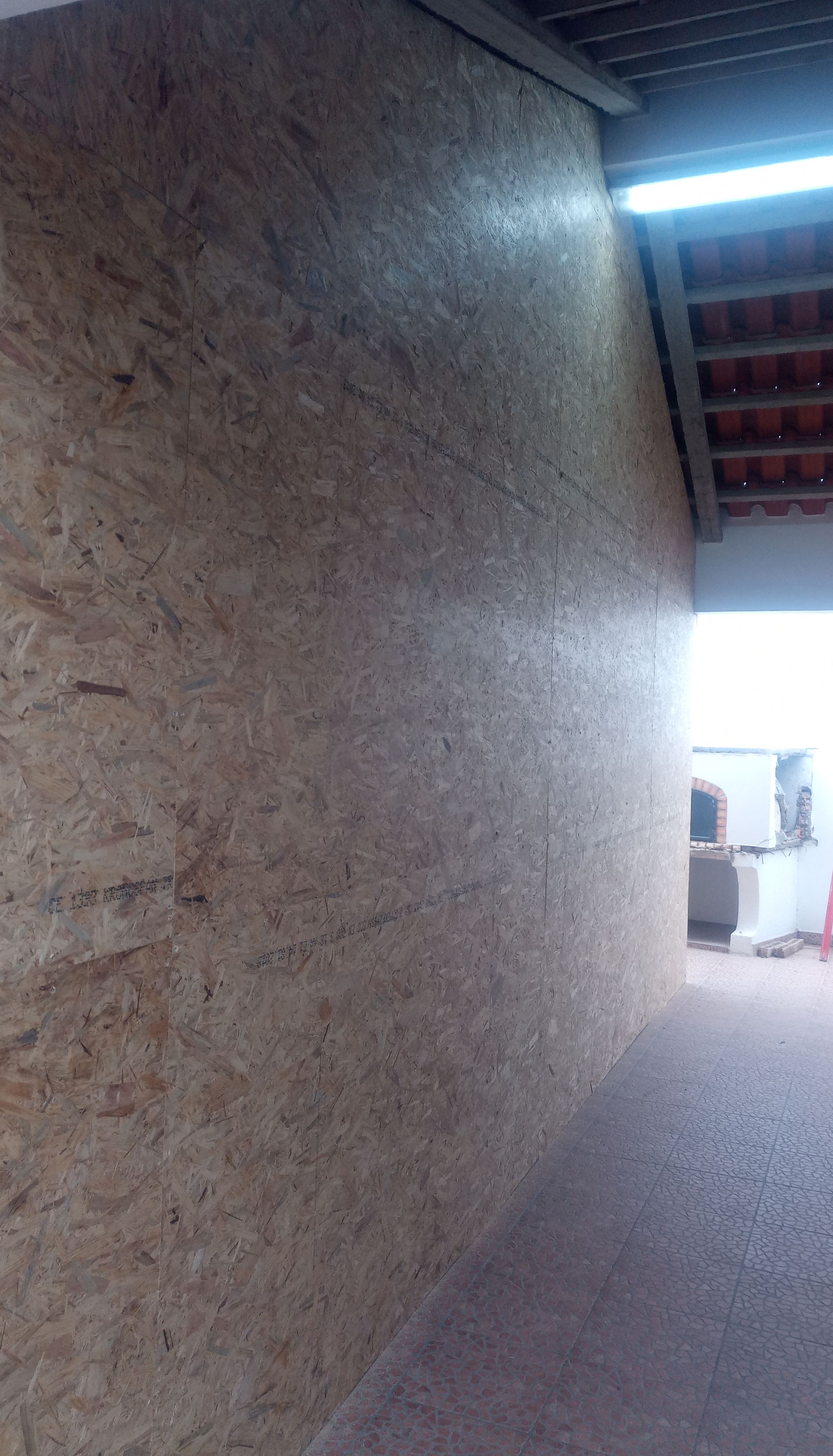 Parede de painéis OSB sob um telhado de metal ondulado. A luz solar entra por uma abertura à direita.