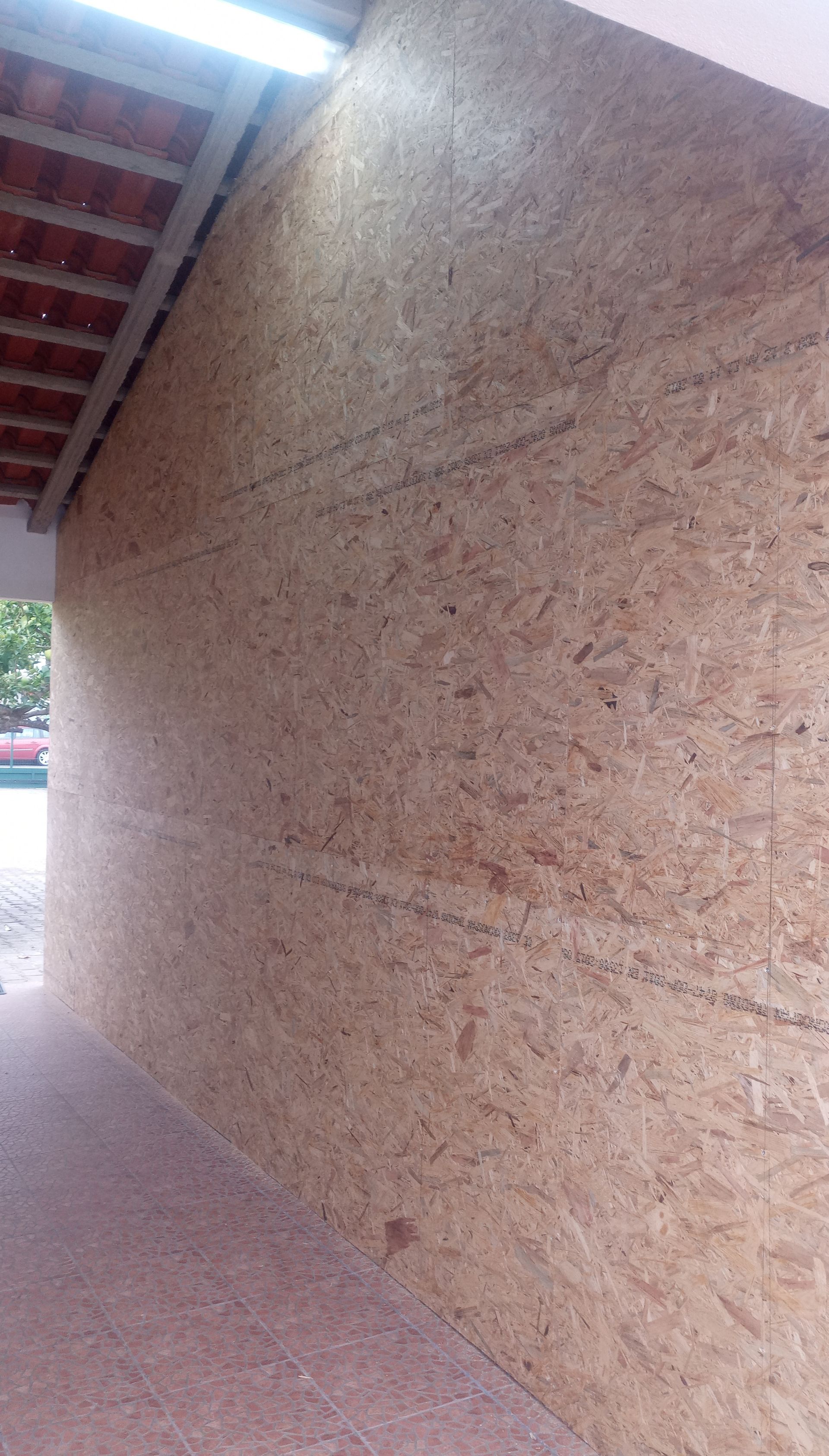 Parede revestida com painéis OSB sob uma área coberta, com um telhado de tijolos vermelhos visível na parte superior.