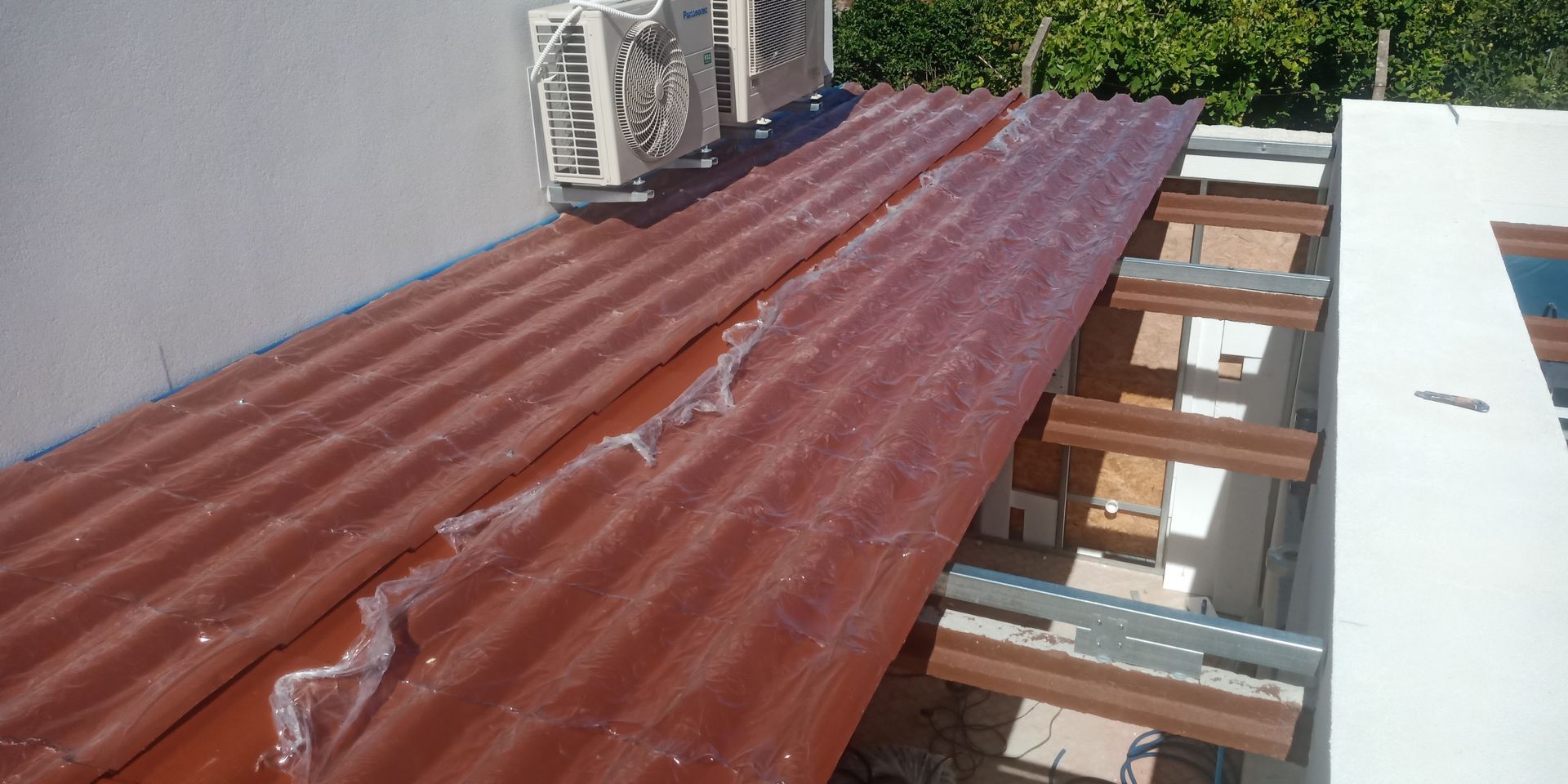 Telhado de metal ondulado vermelho, com cobertura plástica, estende-se da parede. Vigas de madeira e unidades de ar condicionado são visíveis.