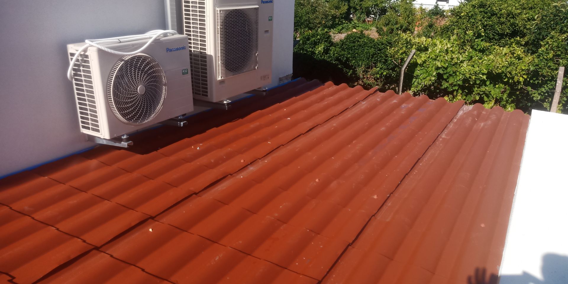 Duas unidades de ar condicionado em um telhado de metal ondulado vermelho, com folhagem verde ao fundo.