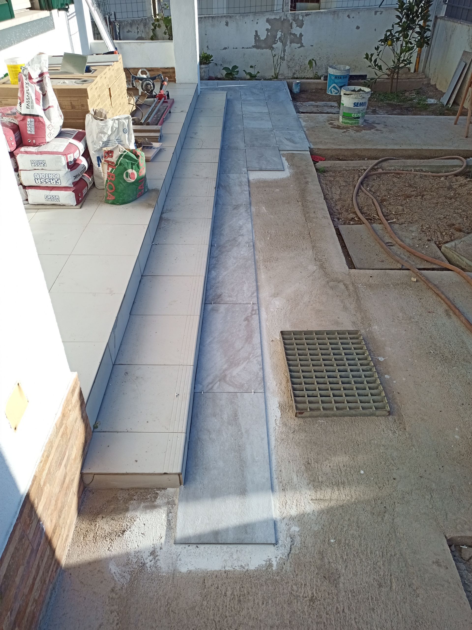Rampa externa em construção com concreto e azulejo. Abastecimento e grelha de drenagem visíveis.