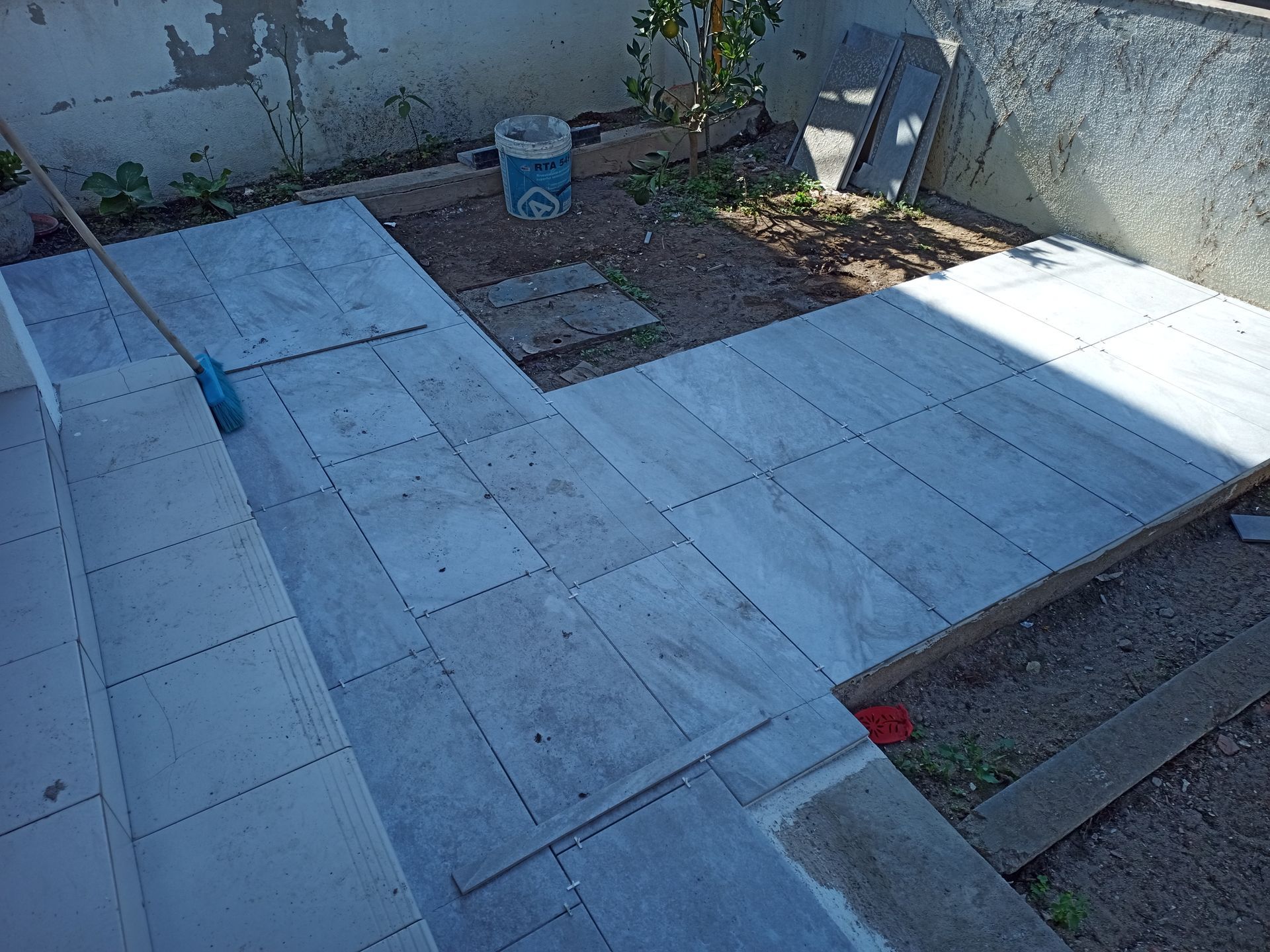 Caminho em construção, ladrilhos claros sendo assentados em um pátio, degraus de concreto à esquerda, chão de terra.