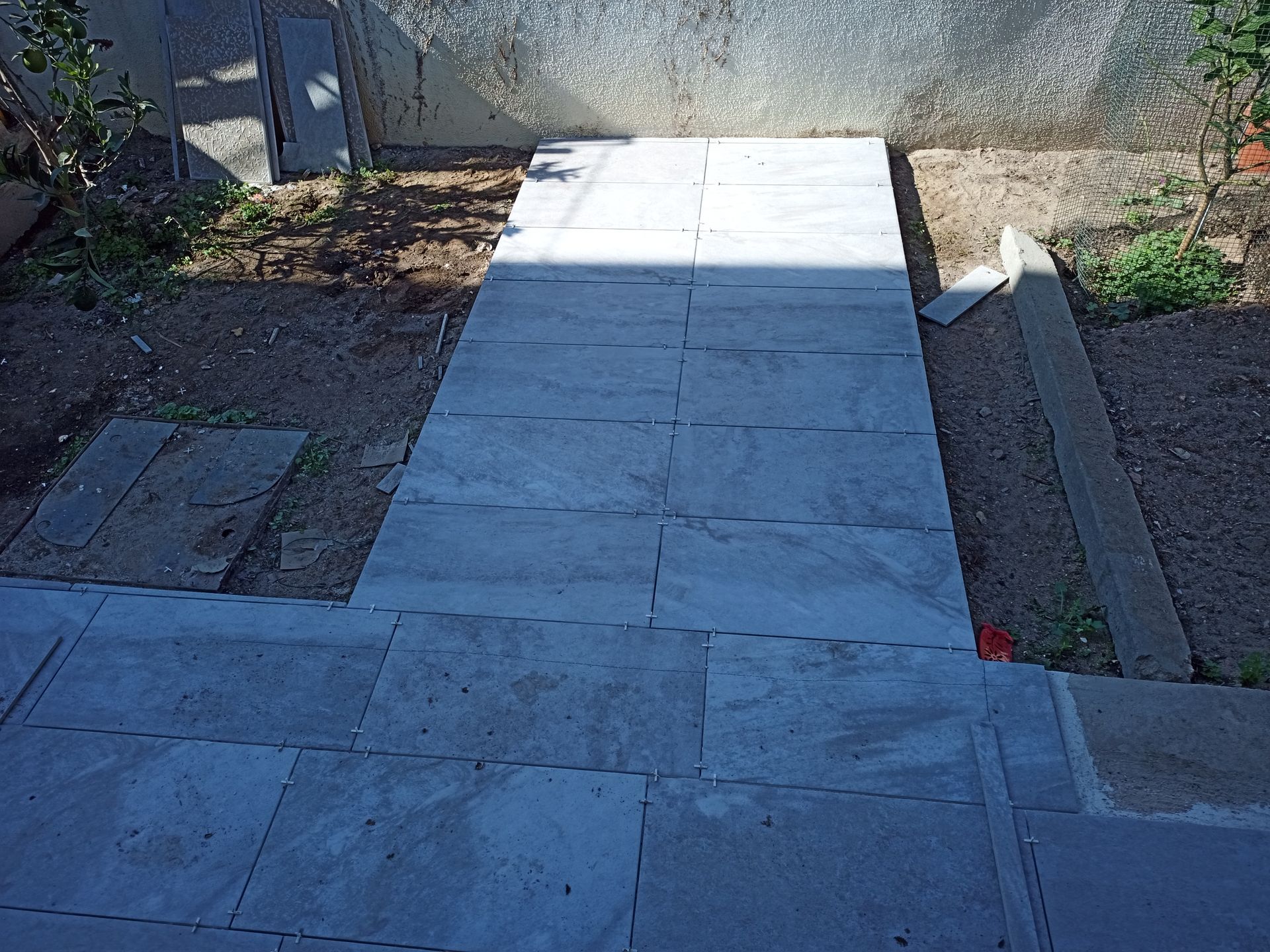 Caminho de pedra que parte de um pátio, rodeado de terra e vegetação, e sobe até um muro.