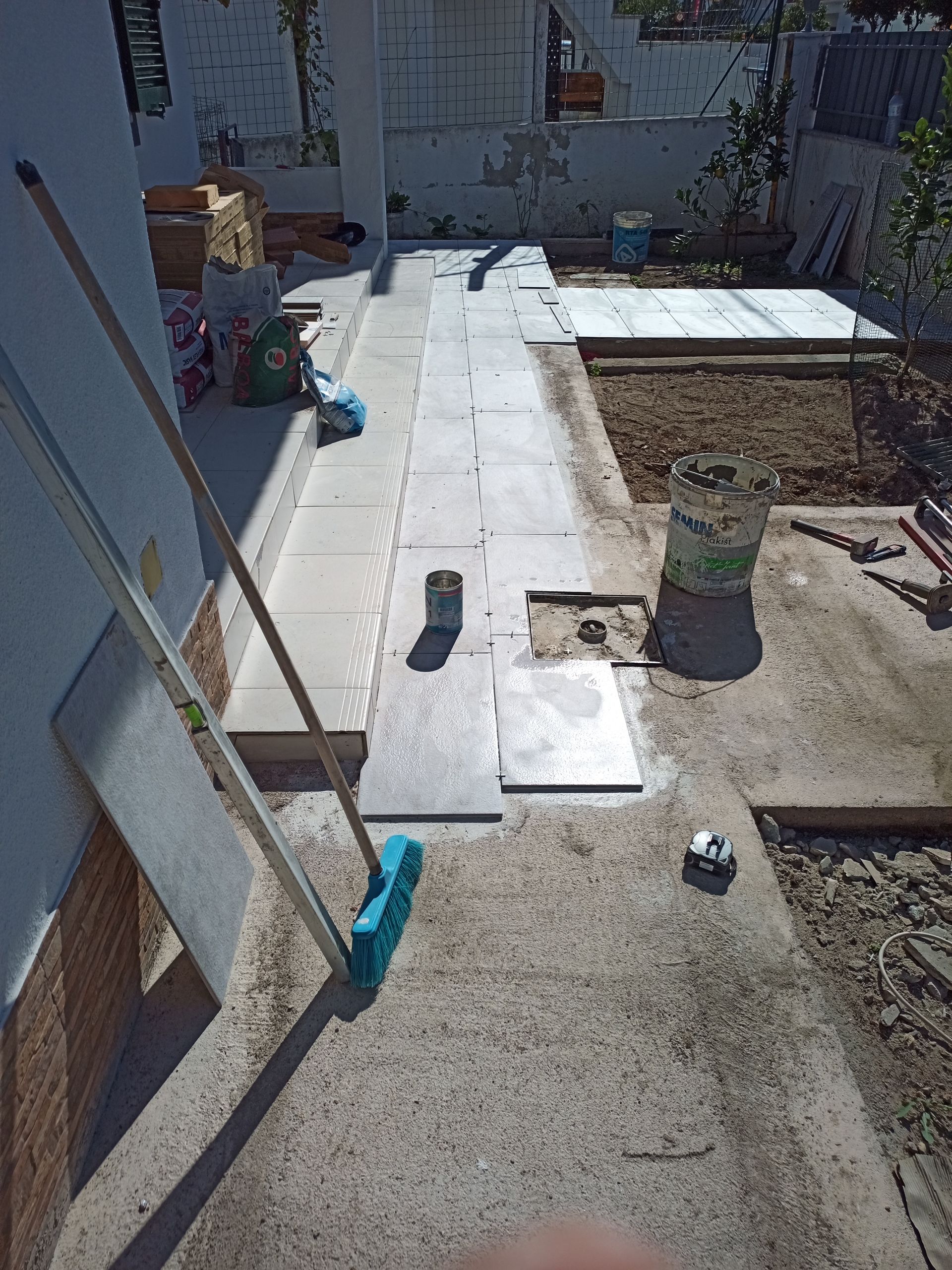 Um pátio em construção com ladrilhos retangulares sendo assentados. Ferramentas e materiais estão espalhados pela área de trabalho.