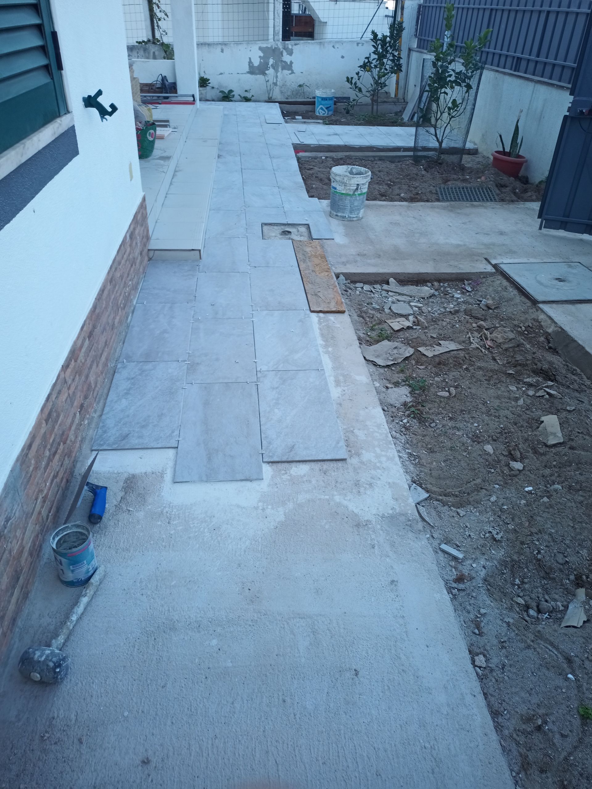 Construção de calçada: Ladrilhos de pedra cinza sendo assentados sobre um caminho de concreto. Baldes e ferramentas são visíveis.