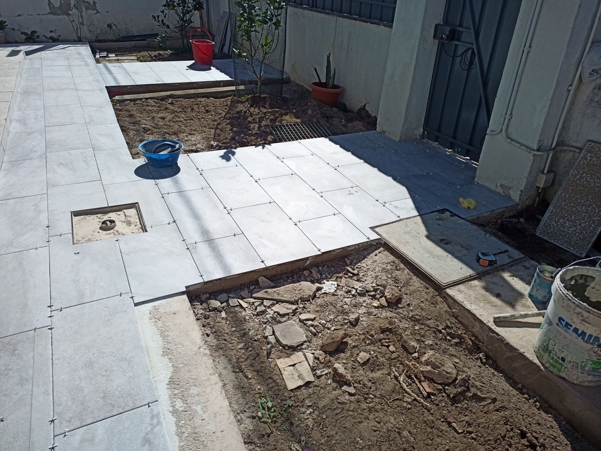 Construção de calçada: ladrilhos cinzentos assentados em um quintal, parcialmente concluído, com canteiros e baldes.