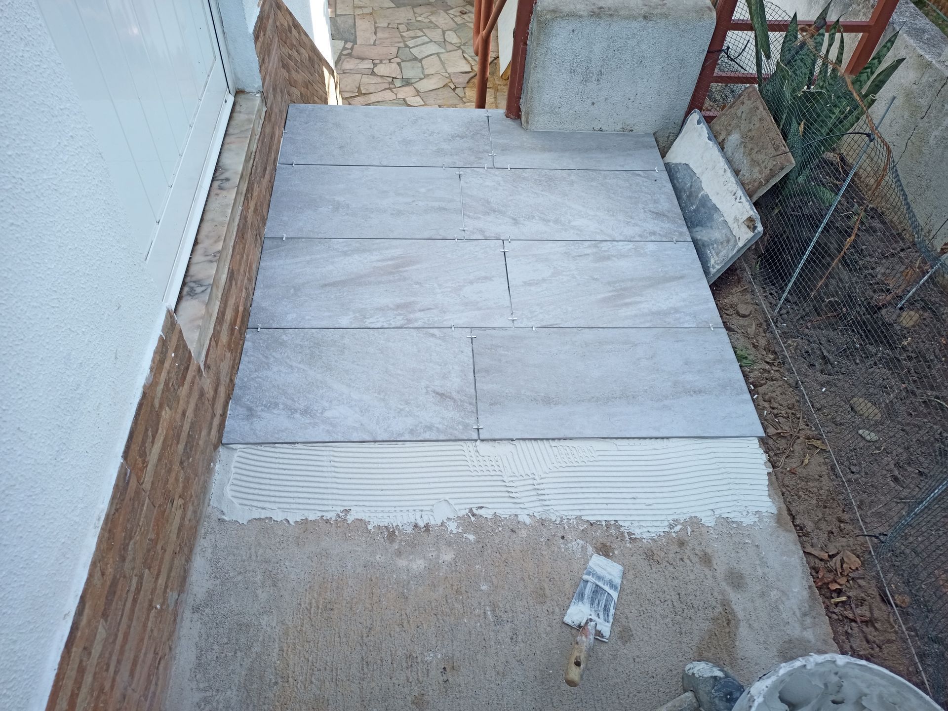 Azulejos cinzentos sendo assentados sobre uma superfície de concreto externa, com ferramentas e materiais visíveis.