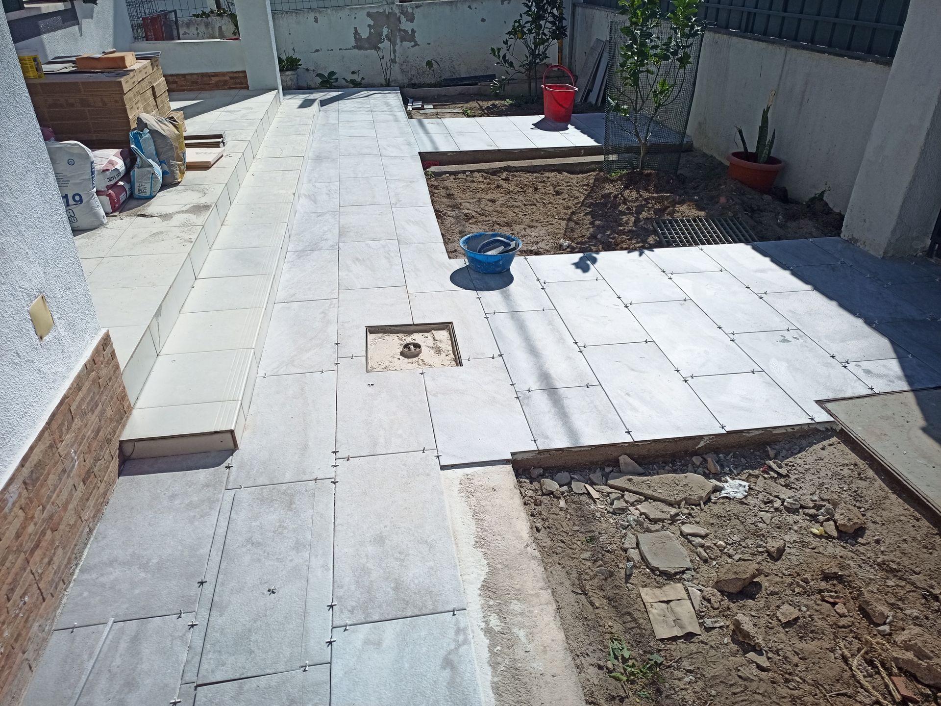 Pátio nos fundos em construção com lajes de pedra. É possível ver terra, ferramentas e parte da parede de tijolos.