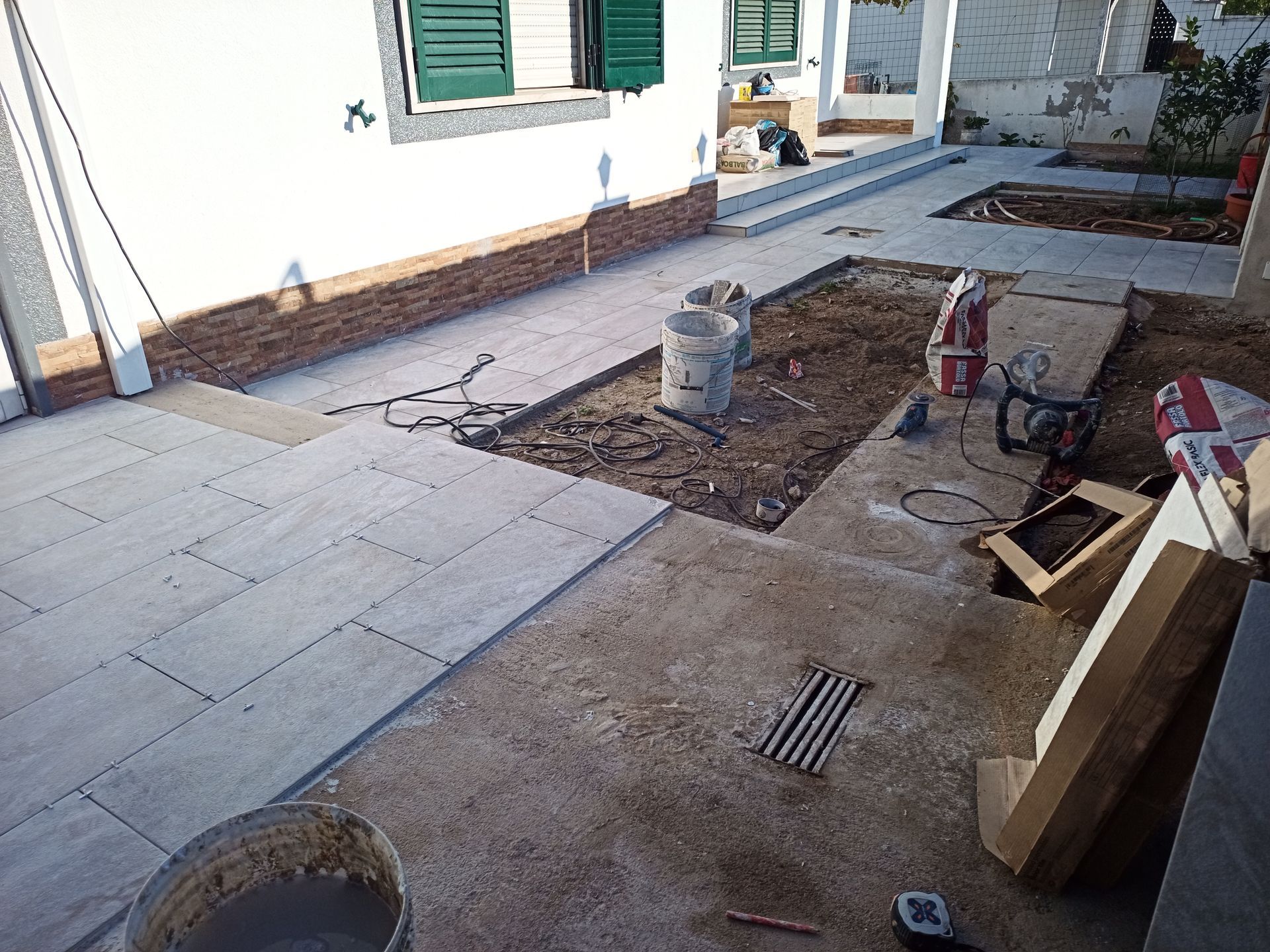 Canteiro de obras com colocação de paralelepípedos, balde de argamassa e materiais de construção perto de uma casa branca.