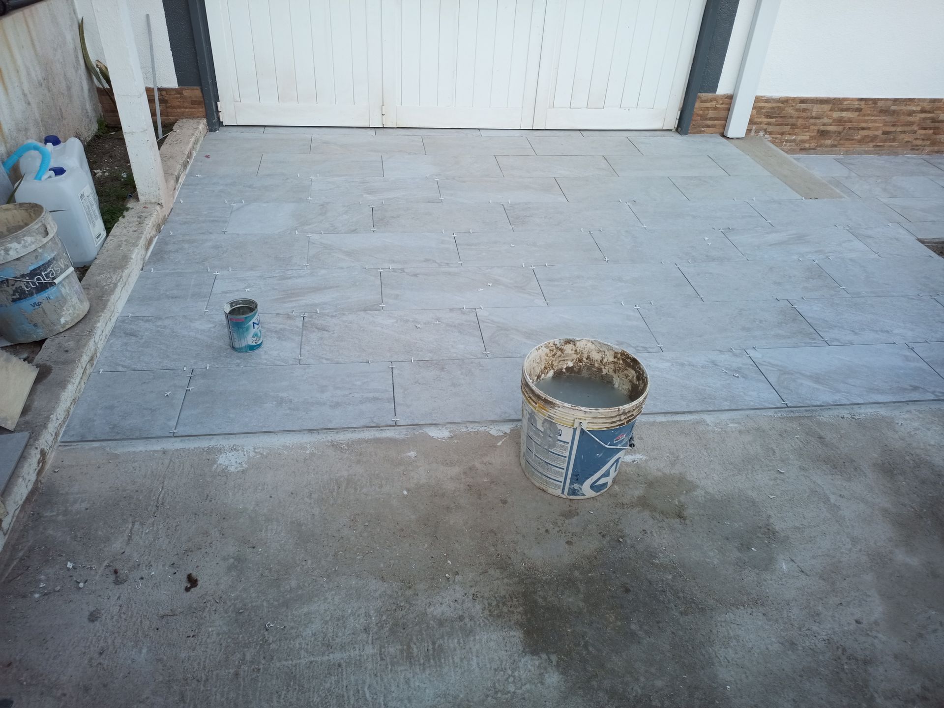 Pátio de cimento com um balde de cimento, um recipiente e um portão branco ao fundo.