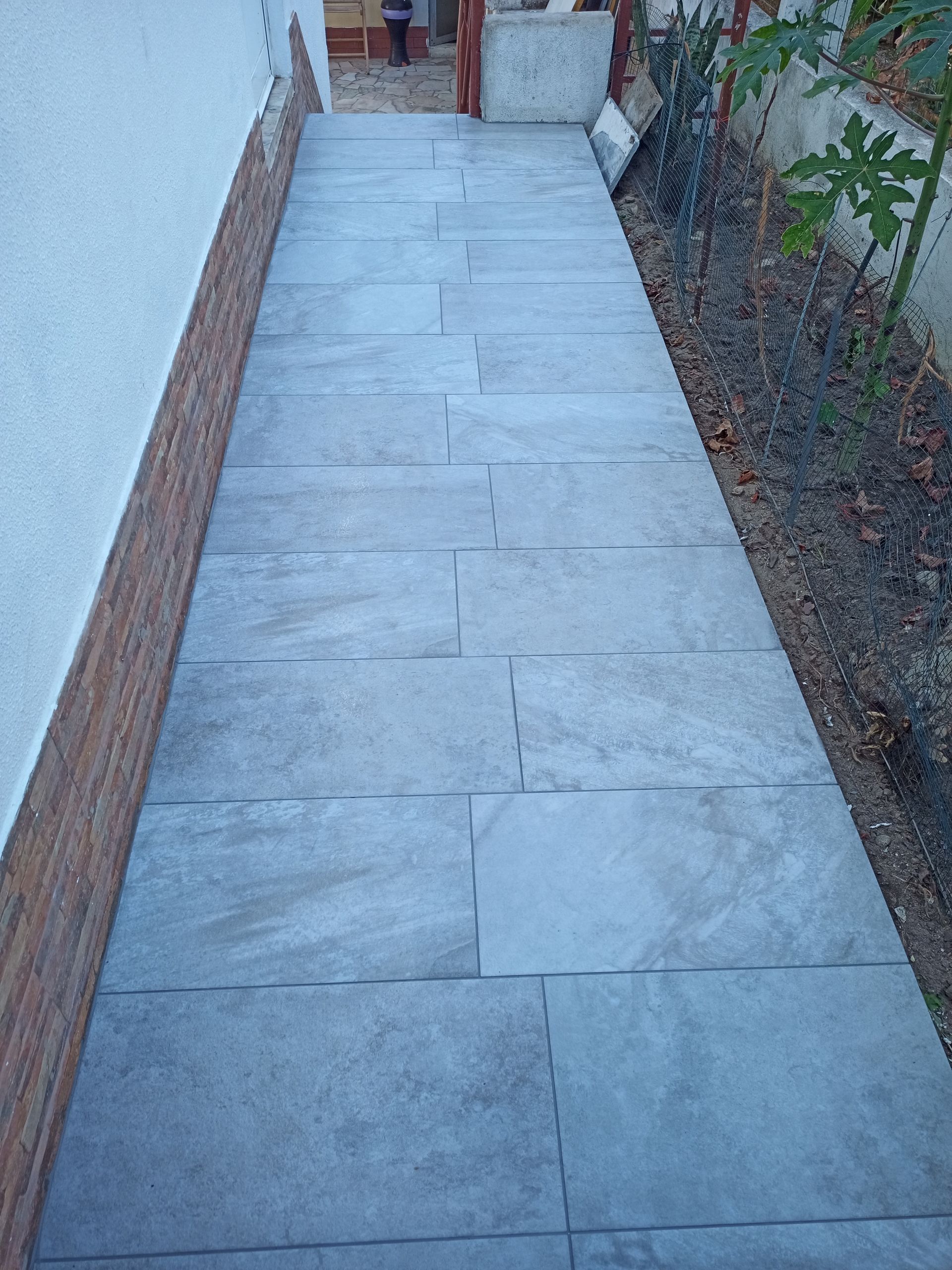 Caminho de ladrilhos cinzentos entre uma parede branca e um canteiro de jardim, que leva a um portão de madeira.