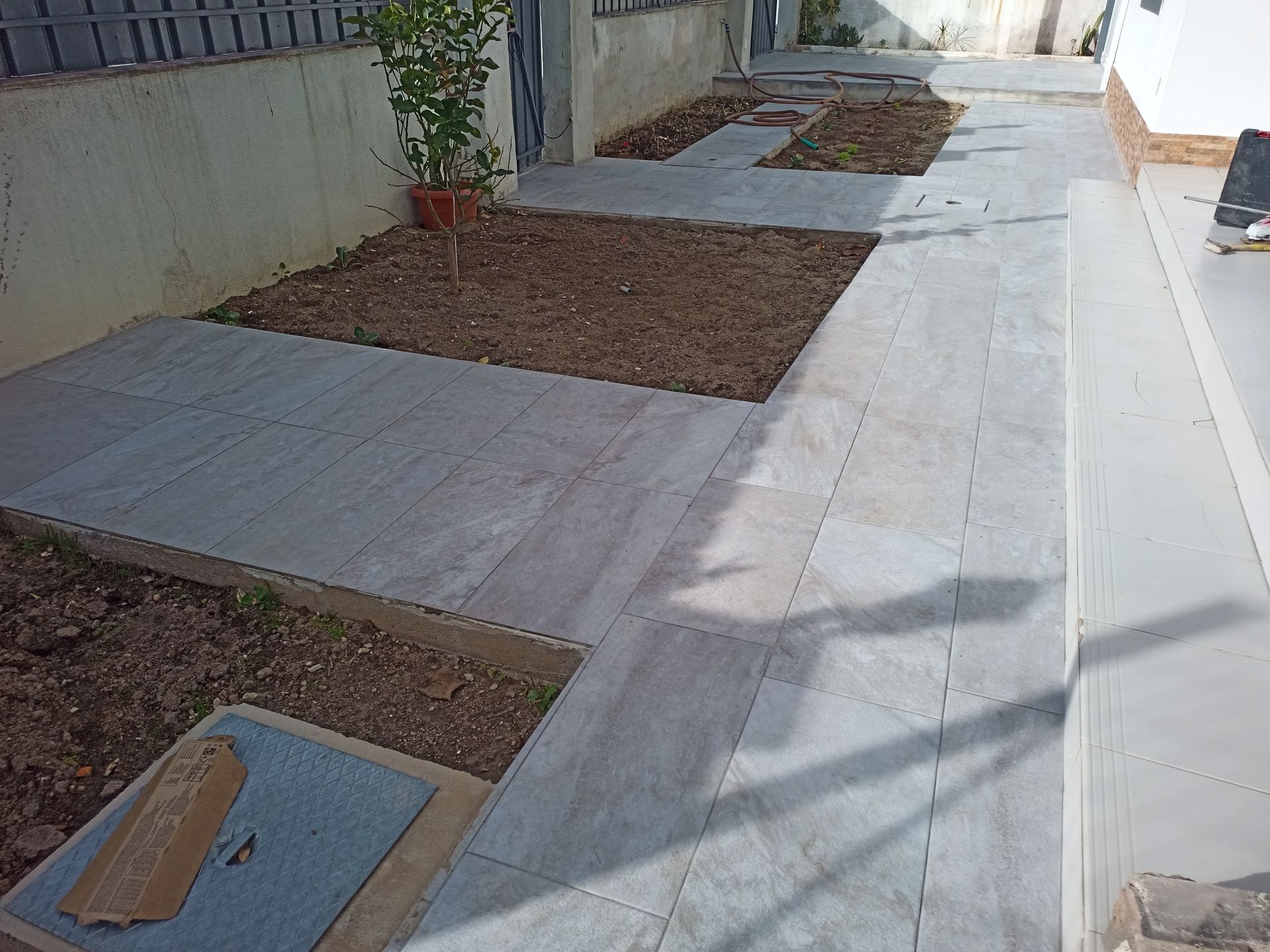 Caminho de pedra com canteiros elevados e uma pequena árvore em vaso.