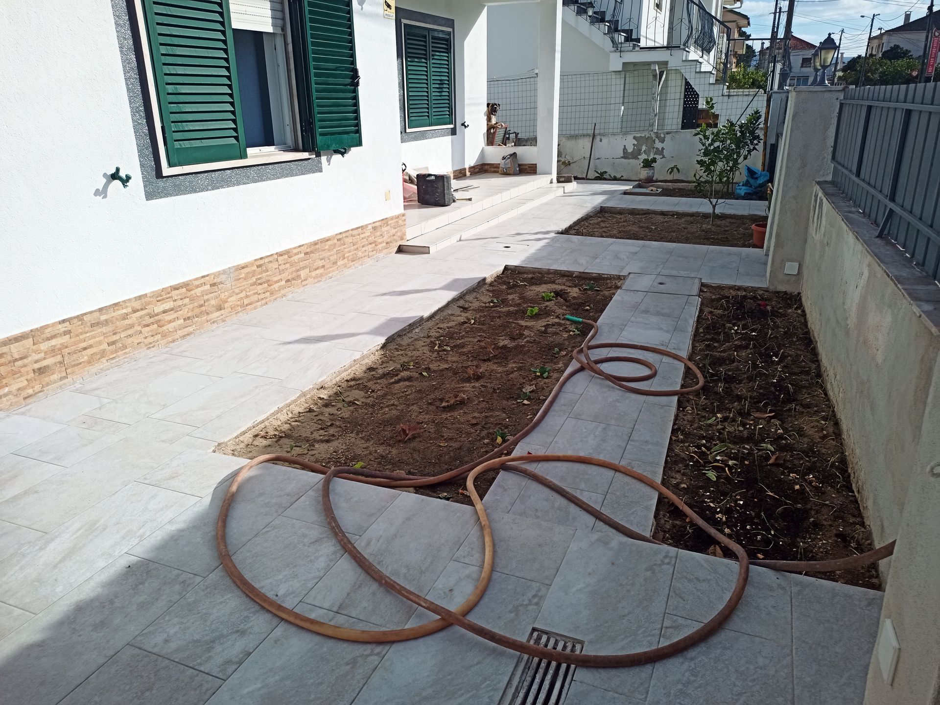 Um caminho com canteiros de flores e uma mangueira ao lado de um prédio branco com venezianas verdes e uma cerca cinza.