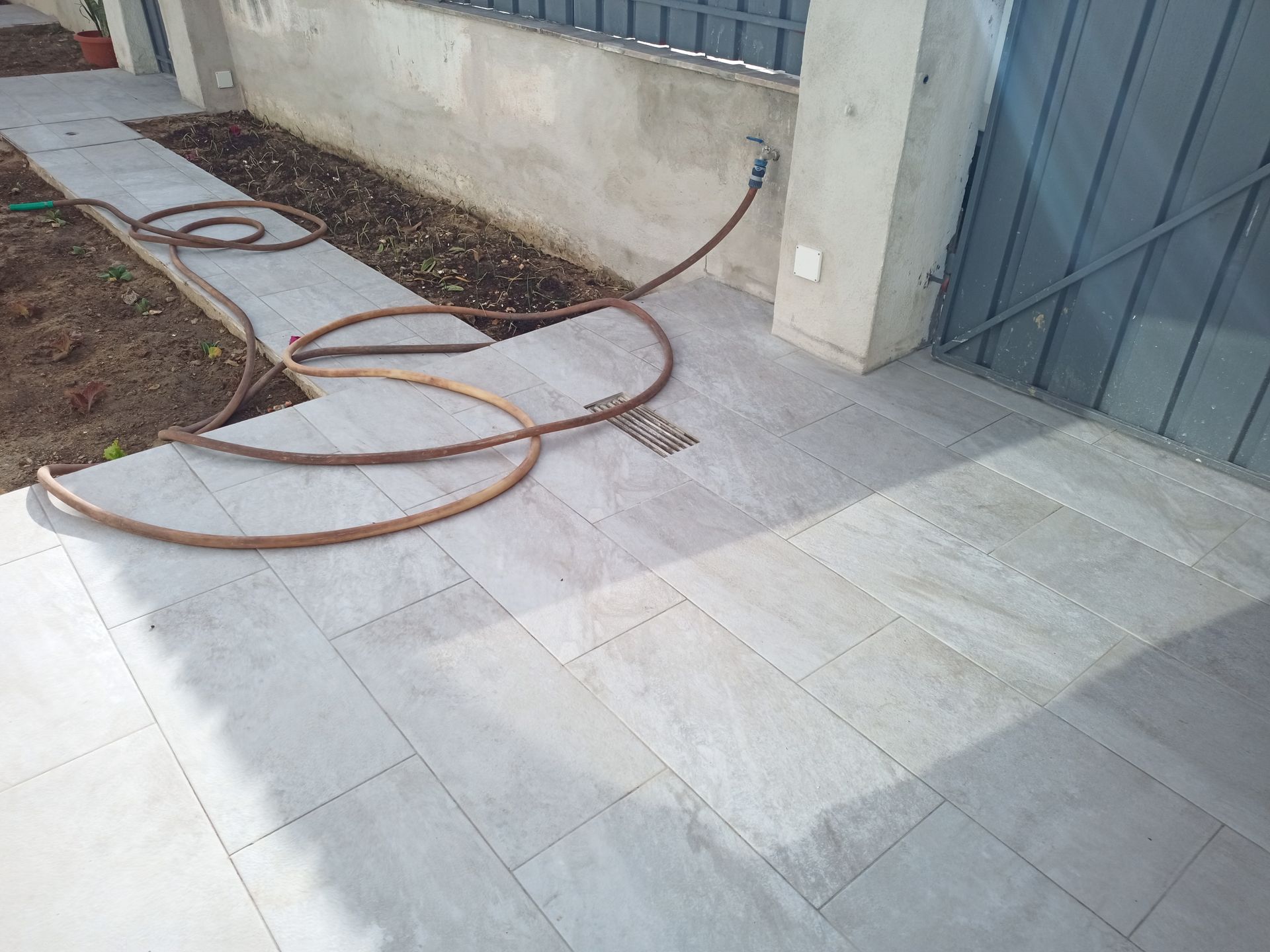 Passarela de ladrilhos cinza com uma mangueira enrolada, que leva a um portão de metal fechado e a um muro de concreto.