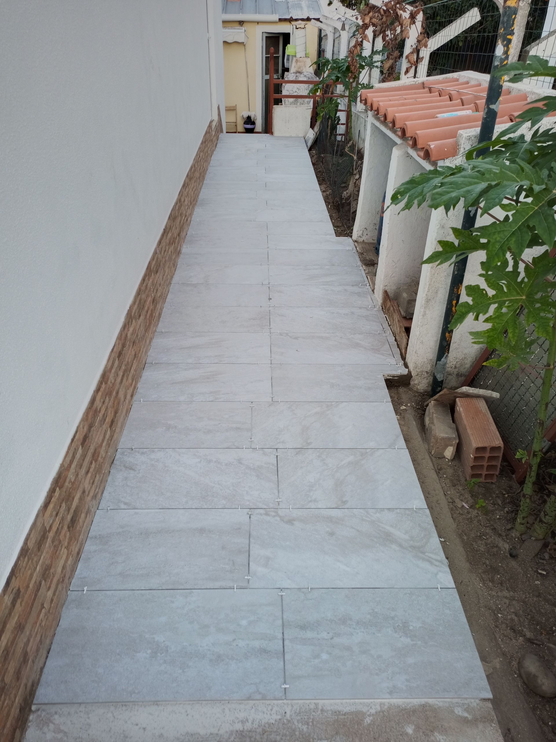 Passagem estreita pavimentada entre uma parede branca e um edifício com telhado de telhas.