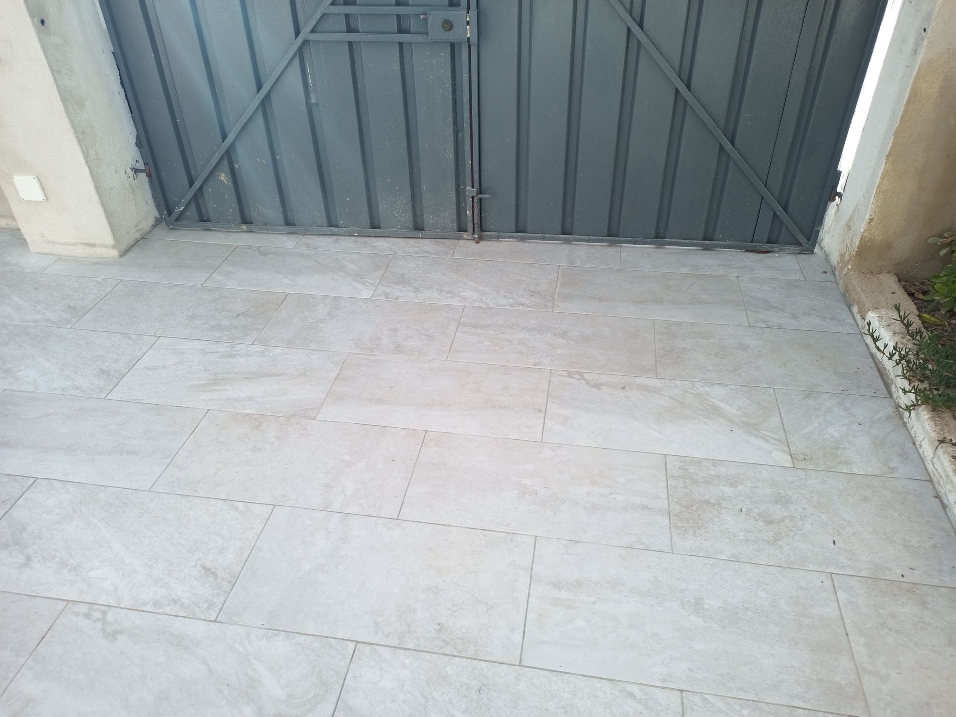 Entrada de automóveis pavimentada com ladrilhos cinzentos que leva a um portão de metal cinzento fechado.