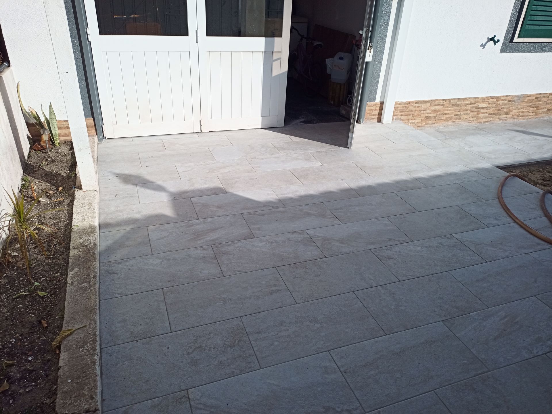 Pátio com azulejos cinza em frente a uma porta de garagem branca, aberta.