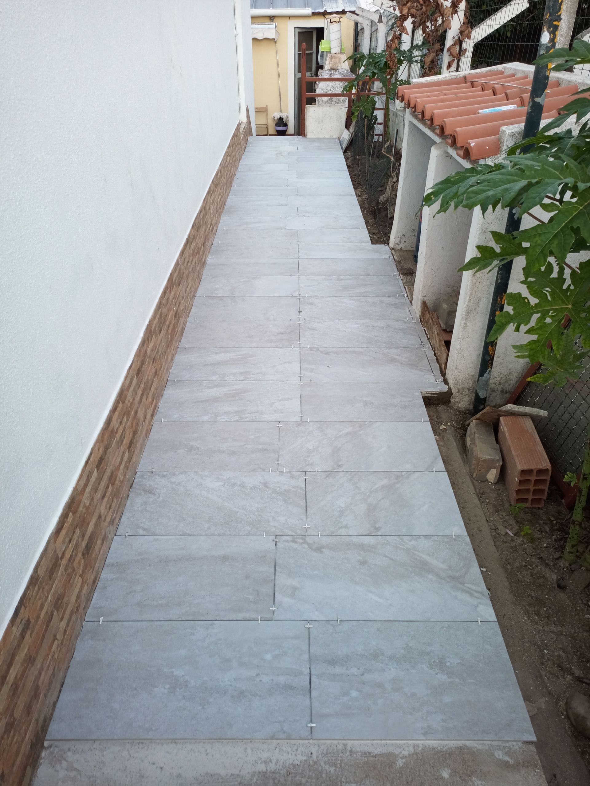 Passarela estreita e pavimentada entre um muro branco e uma área ajardinada.