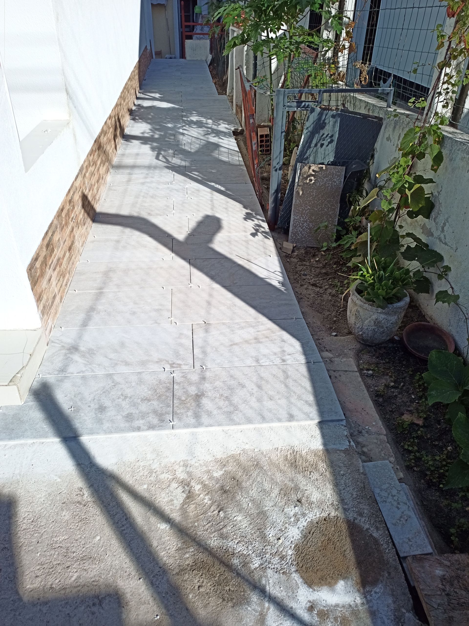 Passarela de concreto com sombra, delimitada por um muro e plantas, que leva a um edifício.