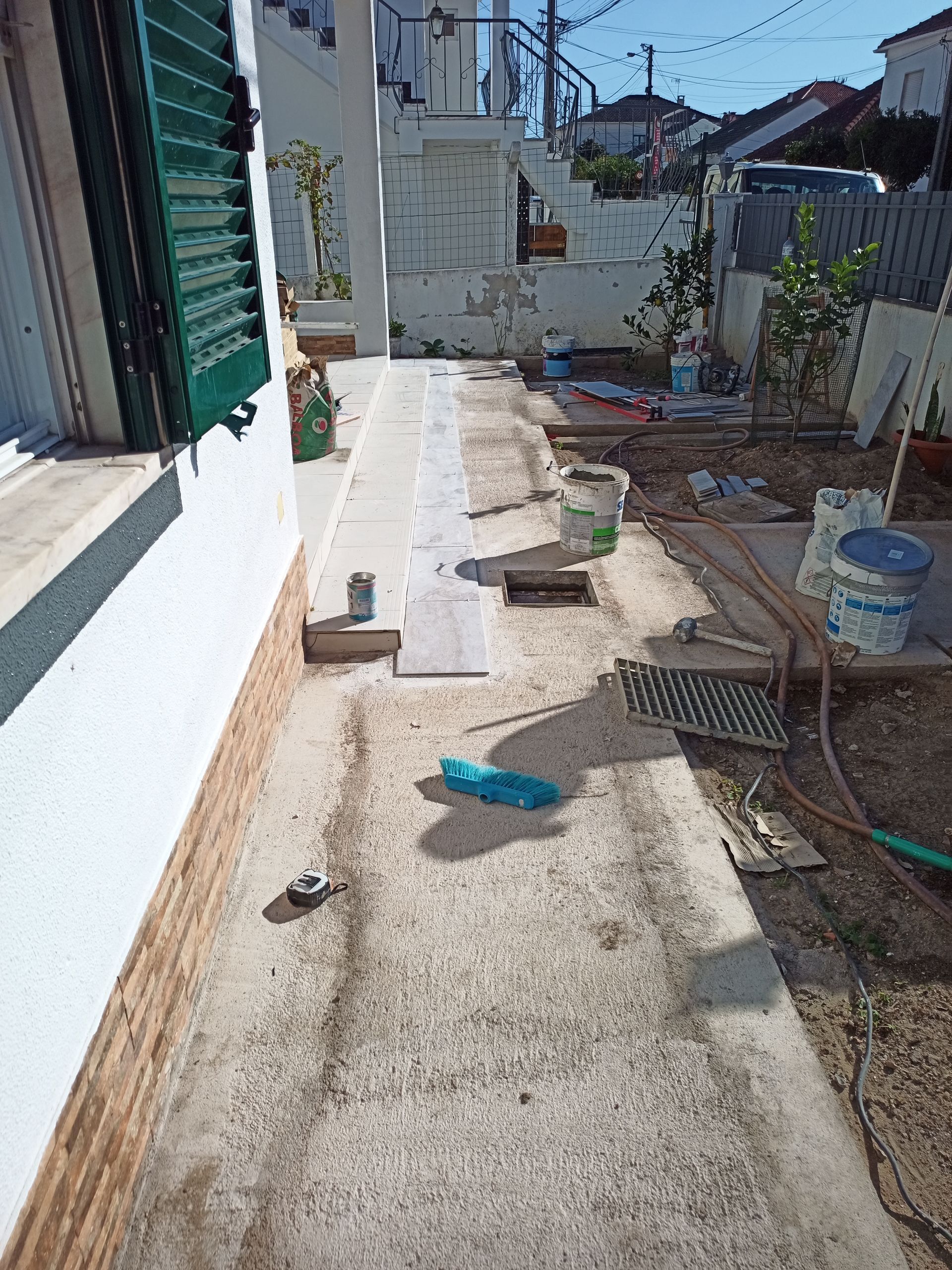 Calçada de concreto ao lado de um prédio branco. Materiais de construção e ferramentas estão espalhados pelo local.