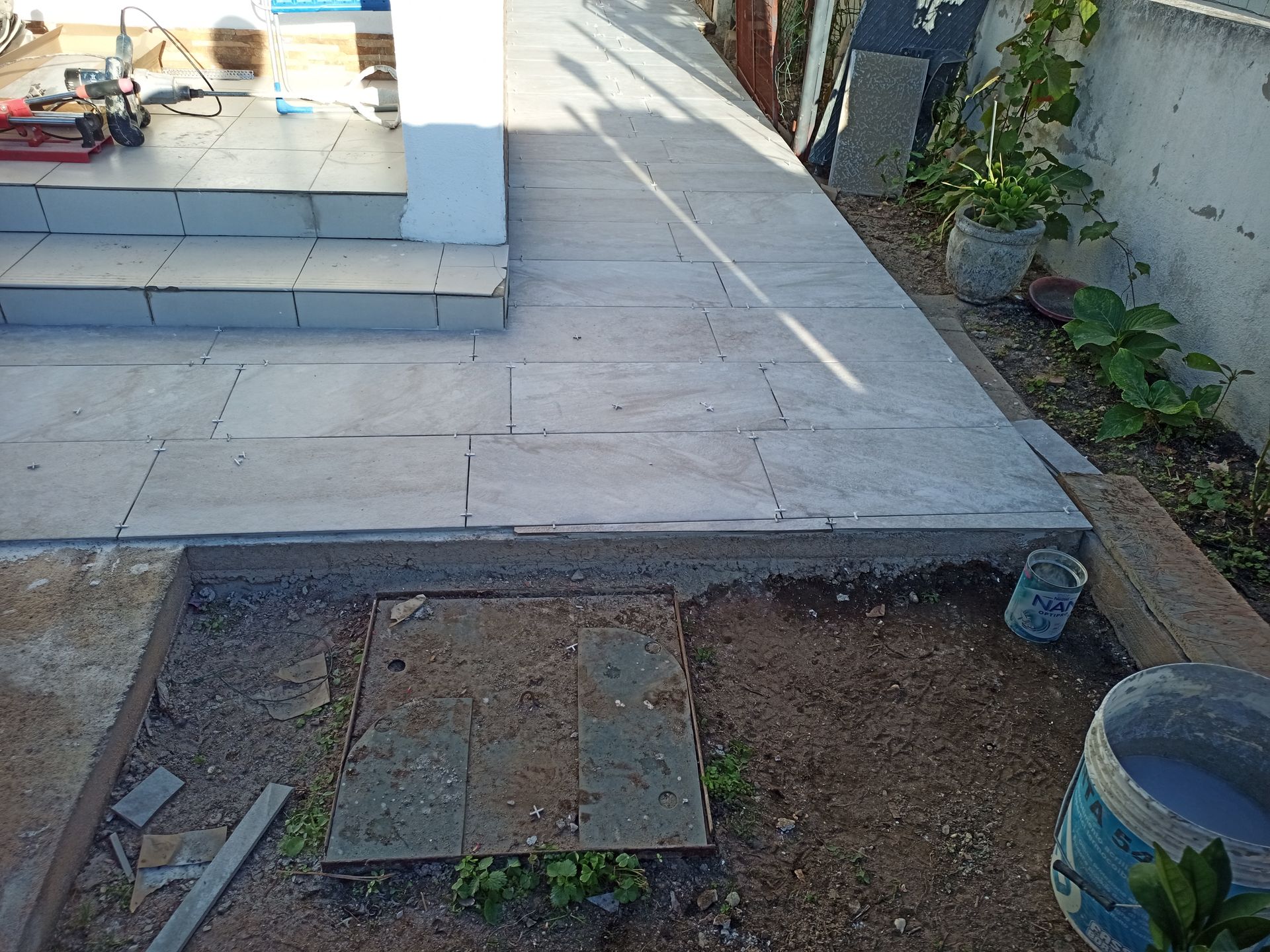 Passarela de ladrilhos cinza com degraus que levam a um prédio e terreno não pavimentado com plantas e suprimentos.